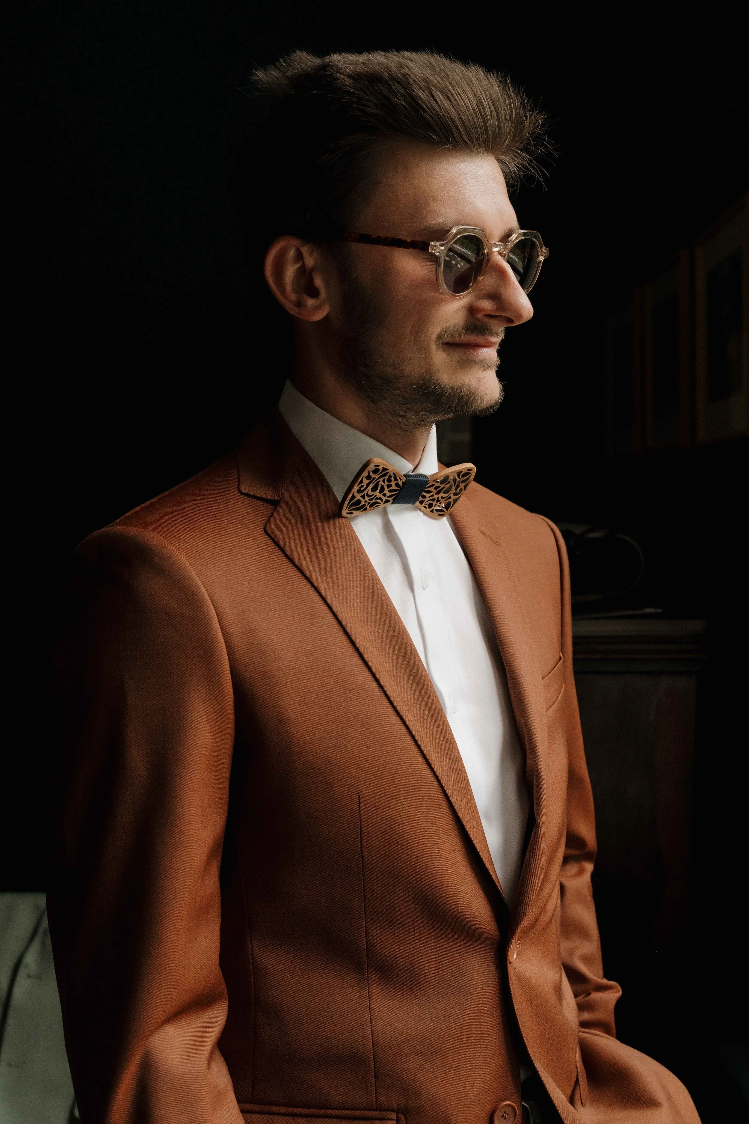Un homme portant un costume marron, des lunettes de soleil, et un nœud papillon en bois, en profil, dans un environnement intérieur Domaine de tourieux à Savigny dans le beaujolais à Lyon. Mariage cérémonie laïque, photographe Lyon haut de gamme, duo