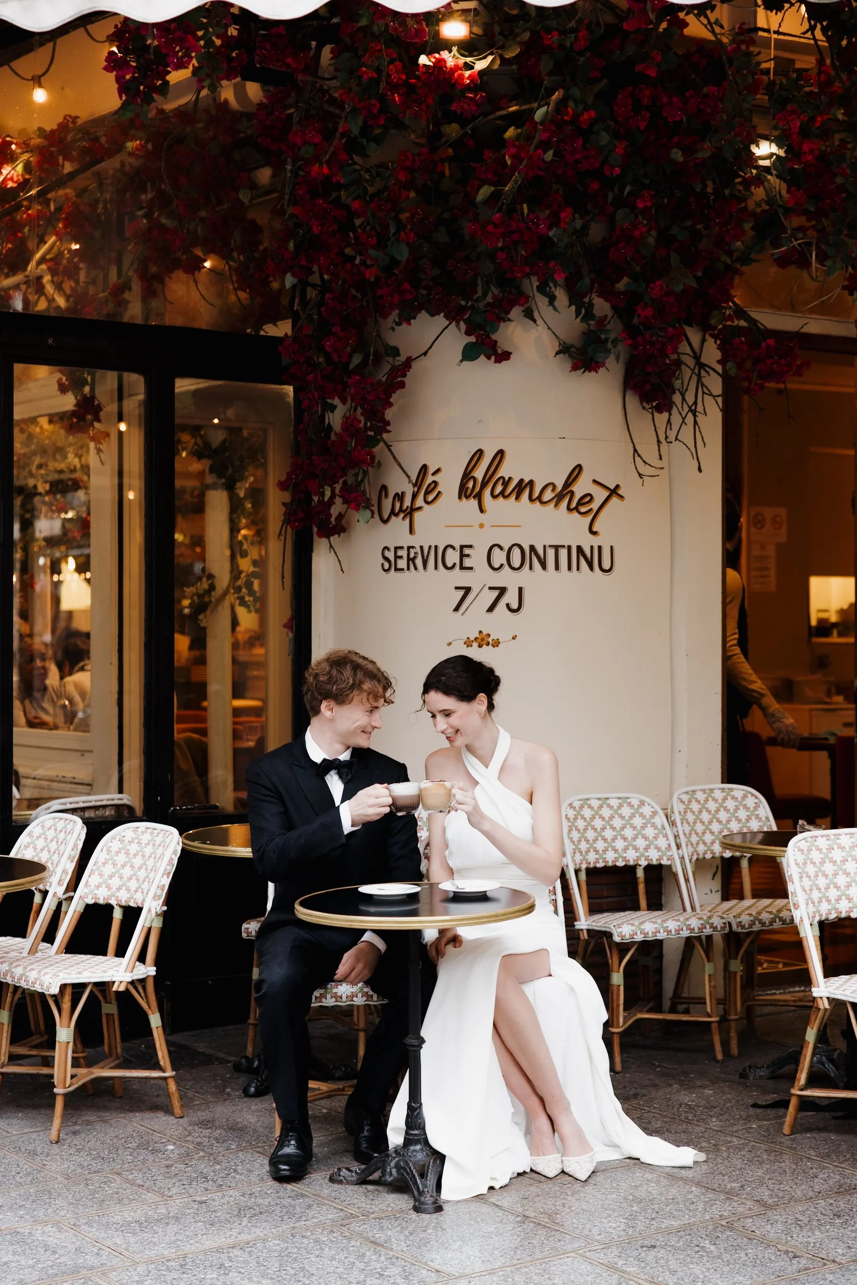 Un couple habillé élégamment, assis à une table extérieure d'un café parisien, partageant un moment de convivialité avec des tasses de café, devant un panneau indiquant le nom du café 'Café Blanchet'. Session engagement café parisien, photographe 