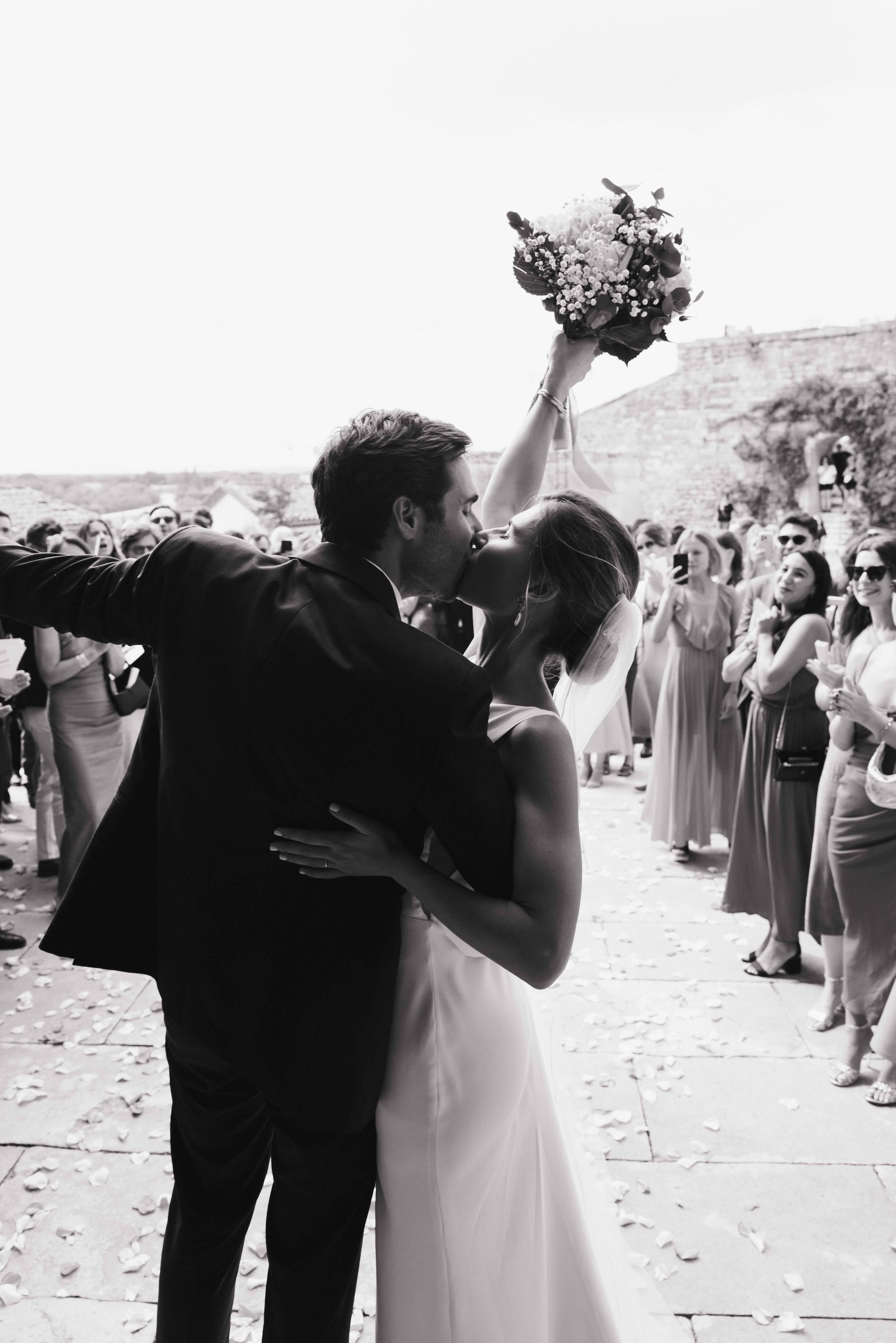 Deux personnes mariées s'embrassent lors de leur mariage, entourées d'invités devant un fond d'architecture ancienne. Domaine du bijoutier à grignan, drome provençal, coucher de soleil, session couple, mariage religieux, cérémonie religieuse 