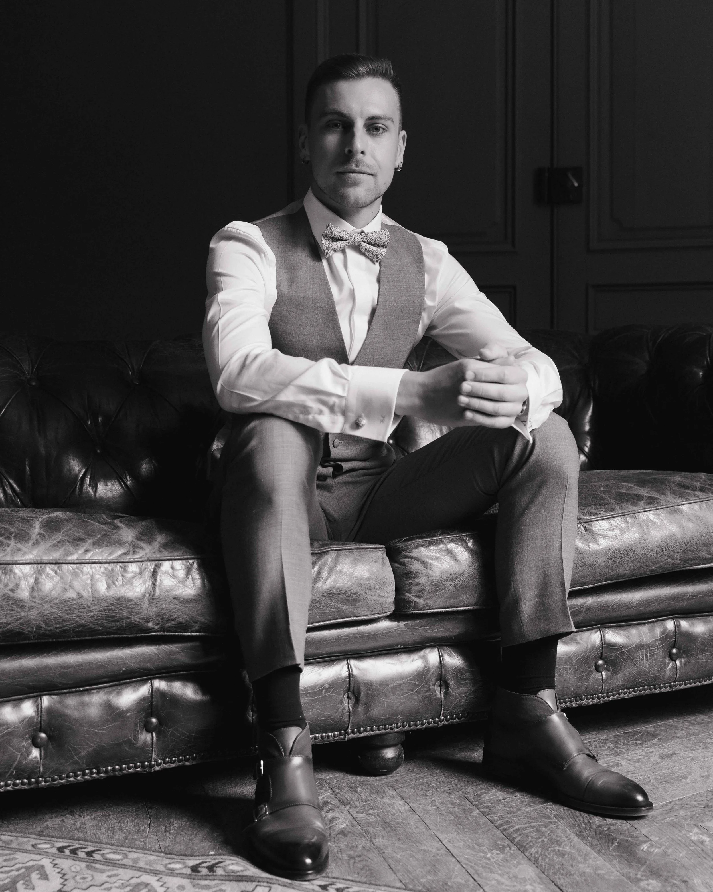 Un homme assis sur un canapé en cuir, habillé en costume gris avec une chemise blanche et un nœud papillon, dans un décor intérieur en noir et blanc. Domaine de tourieux à Savigny dans le beaujolais à Lyon. Mariage cérémonie laïque, photographe Lyon 