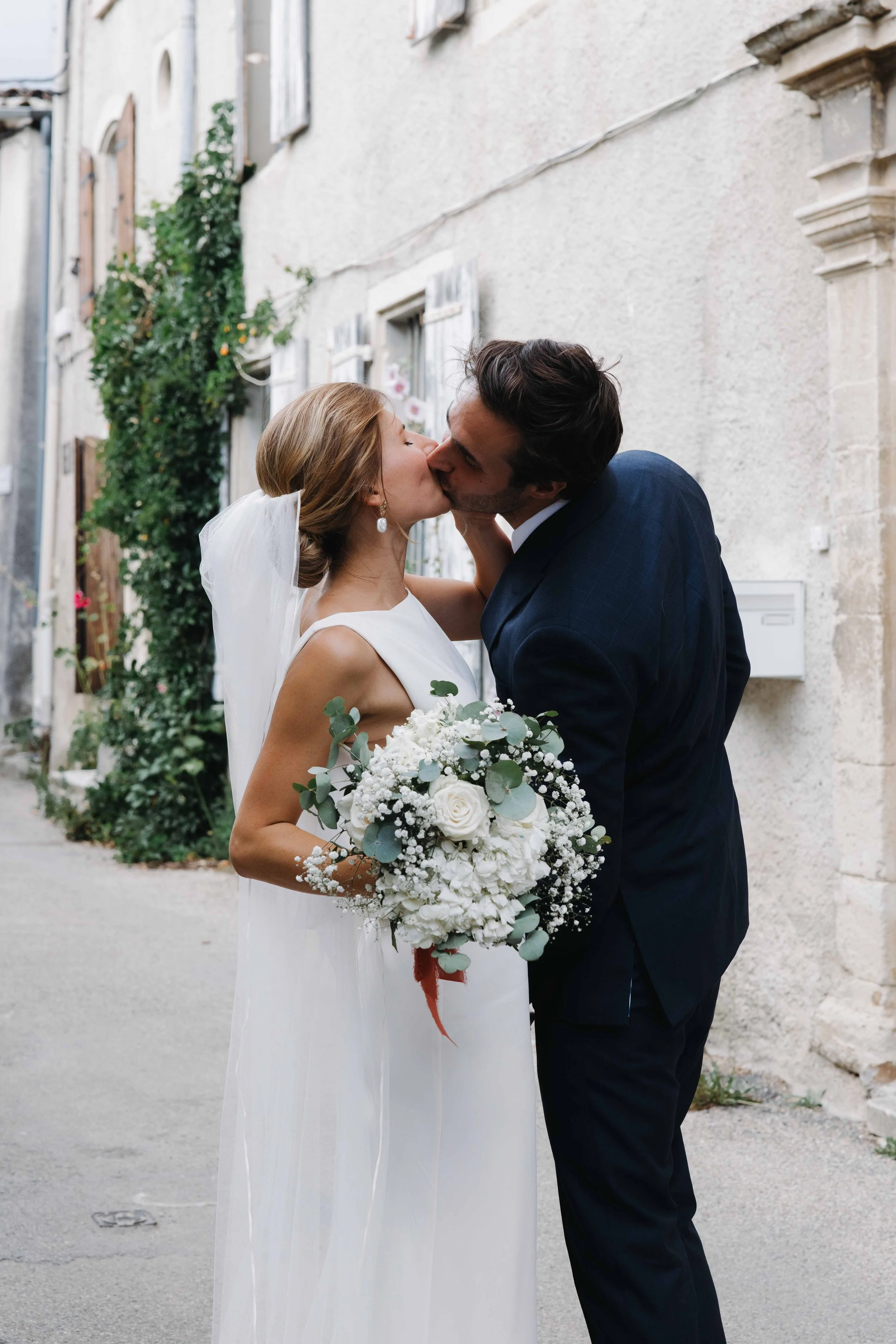Un couple de mariés s'embrasse et échange un baiser lors de leur mariage en plein air, la femme tenant un bouquet de fleurs blanches, dans une rue pavée avec des murs en pierre et des volets en bois. Domaine du bijoutier à grignan, drome provençal 