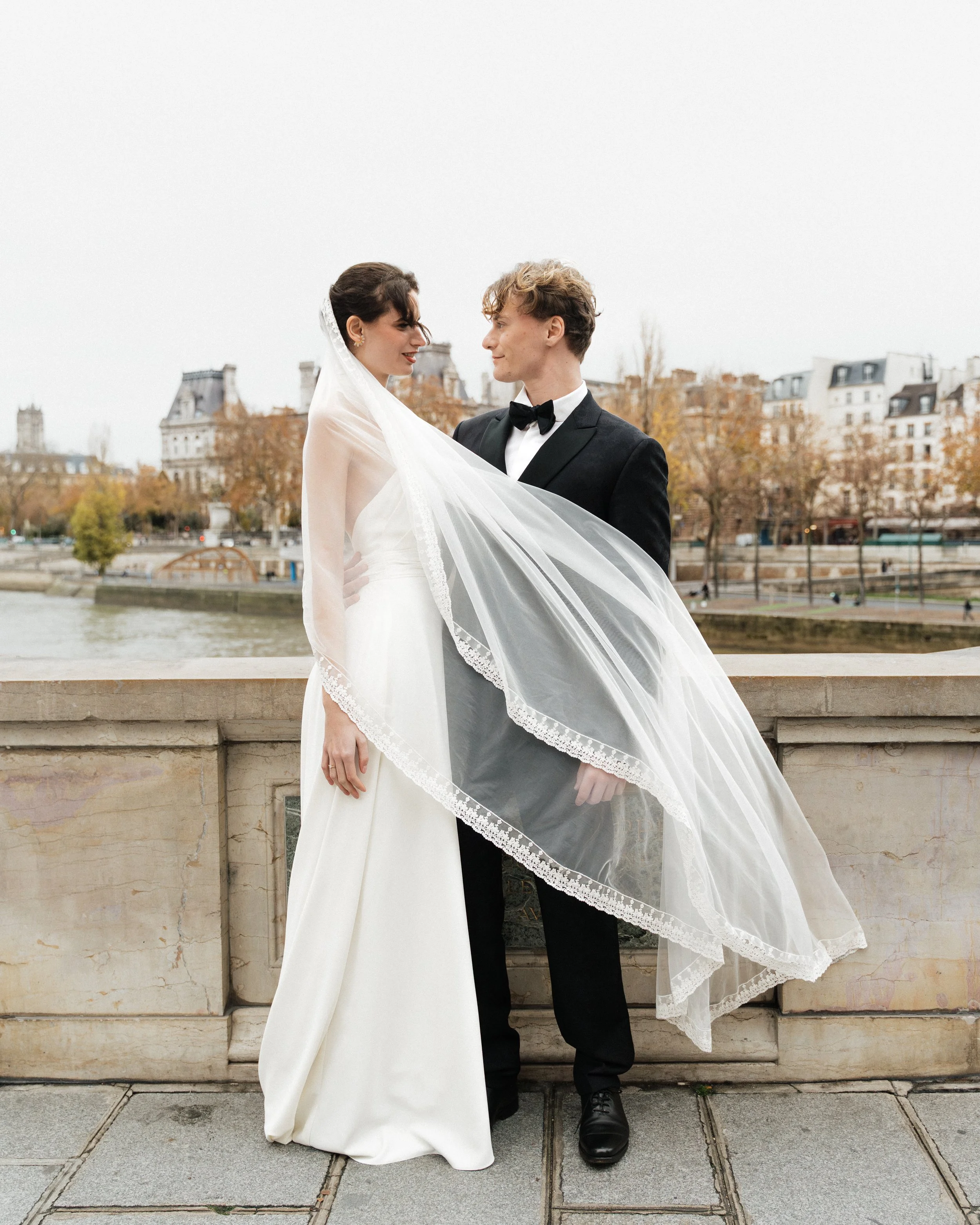 Un couple de mariés, une femme en robe blanche et un homme en costume noir, se regardent avec tendresse sur un pont à Paris.