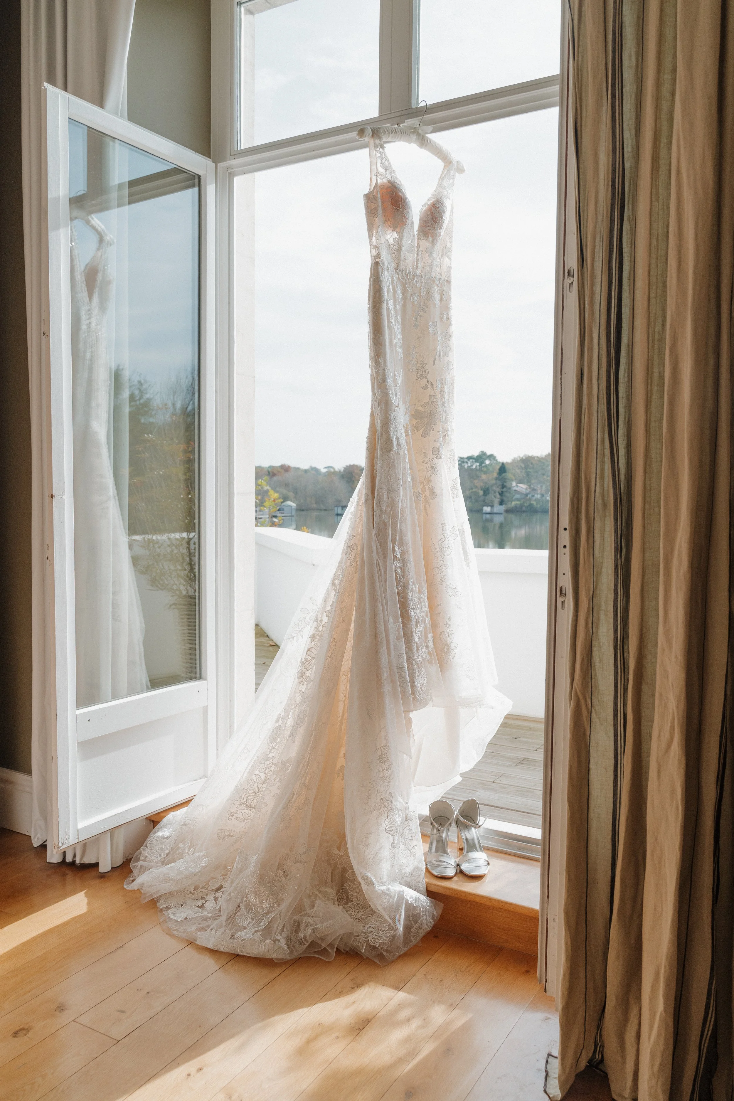 Robe de mariée blanche suspendue à une tringle près d'une fenêtre ouverte, avec une paire de chaussures blanches sous la robe.  session femme mariées et robes haut de gamme, brindos, hôtel et lac,  photographe mariage bordeaux et Lyon et destination 
