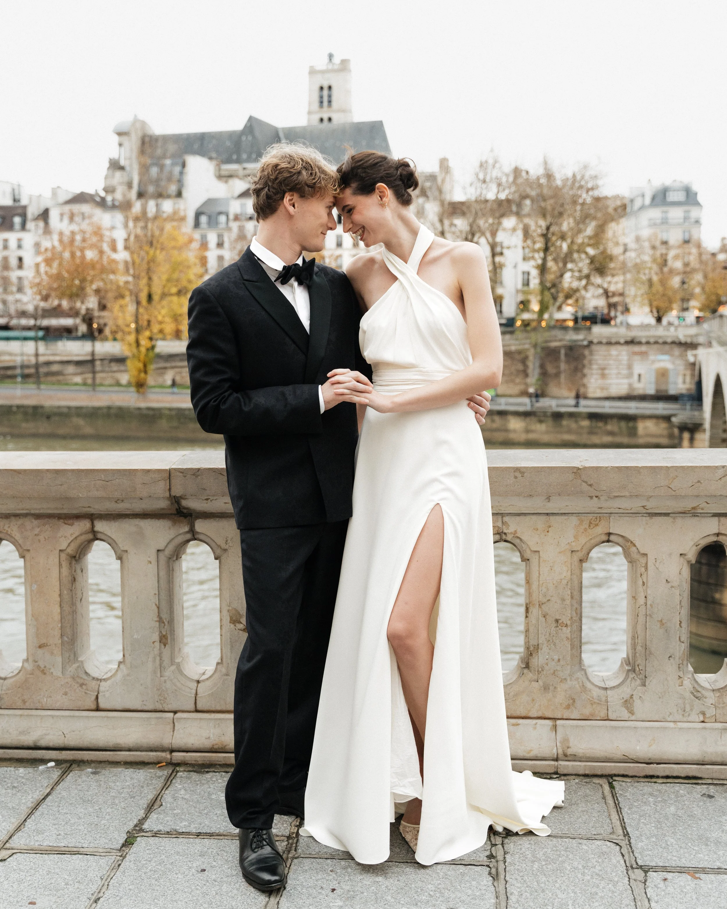 Un couple en vêtements de mariage se tenant la main sur un pont avec un paysage urbain en arrière-plan. Session engagement paris, Louvre, mariage haut de gamme, destination wedding. Robe de mariées, madame a dit oui