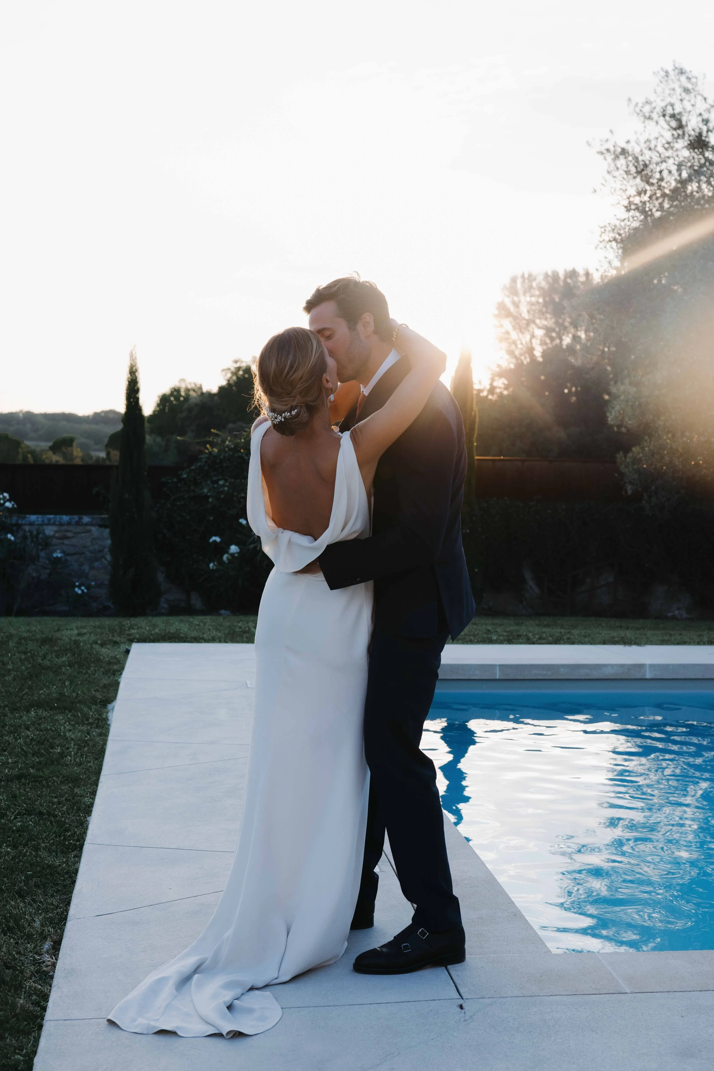 Un couple en robe de mariage et costume s'embrassent près d'une piscine au coucher du soleil. Domaine du bijoutier à grignan, drome provençal, coucher de soleil, session couple, mariage religieux, cérémonie laïque, photographe mariage sud 