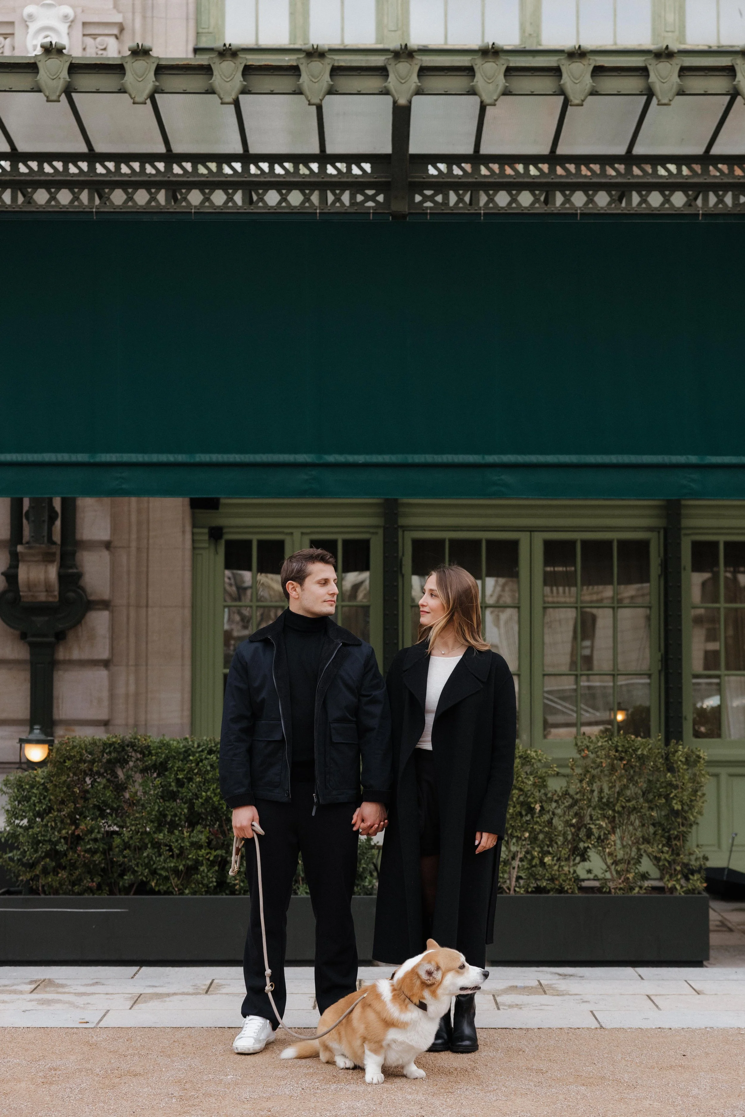 Un couple tenant la main avec leur chien Pembroke Welsh Corgi devant un bâtiment parisien antique avec des fenêtres vertes et des plantes en pots. session engagement couple Lyon 6eme, brotteaux, photographe mariage bordeaux et Lyon et destination