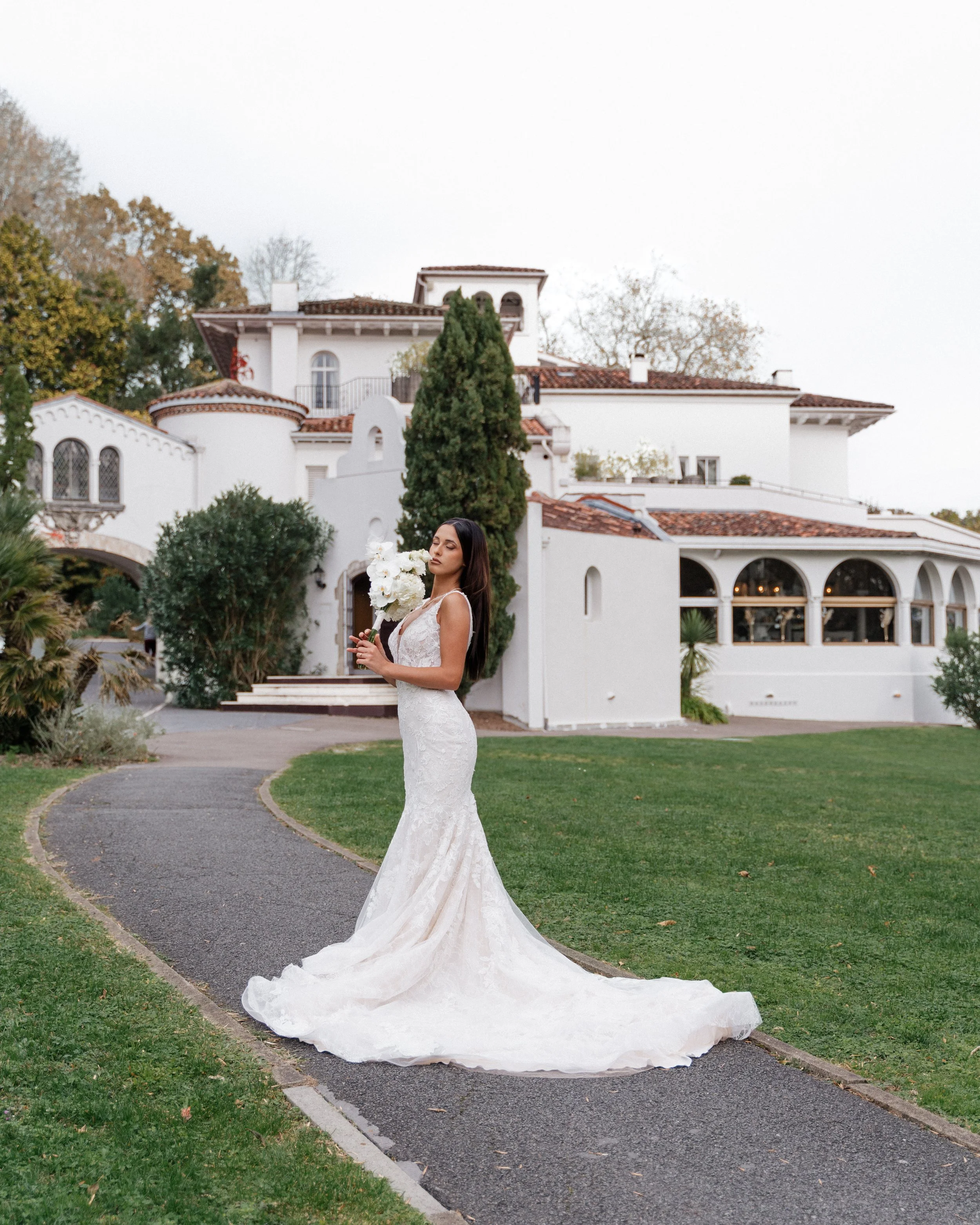 Une femme en robe de mariage blanche tient un bouquet de fleurs blanches devant une grande maison blanche avec jardins et arbres.  session femme mariées et robes haut de gamme, brindos, hôtel et lac,  photographe mariage bordeaux et Lyon 