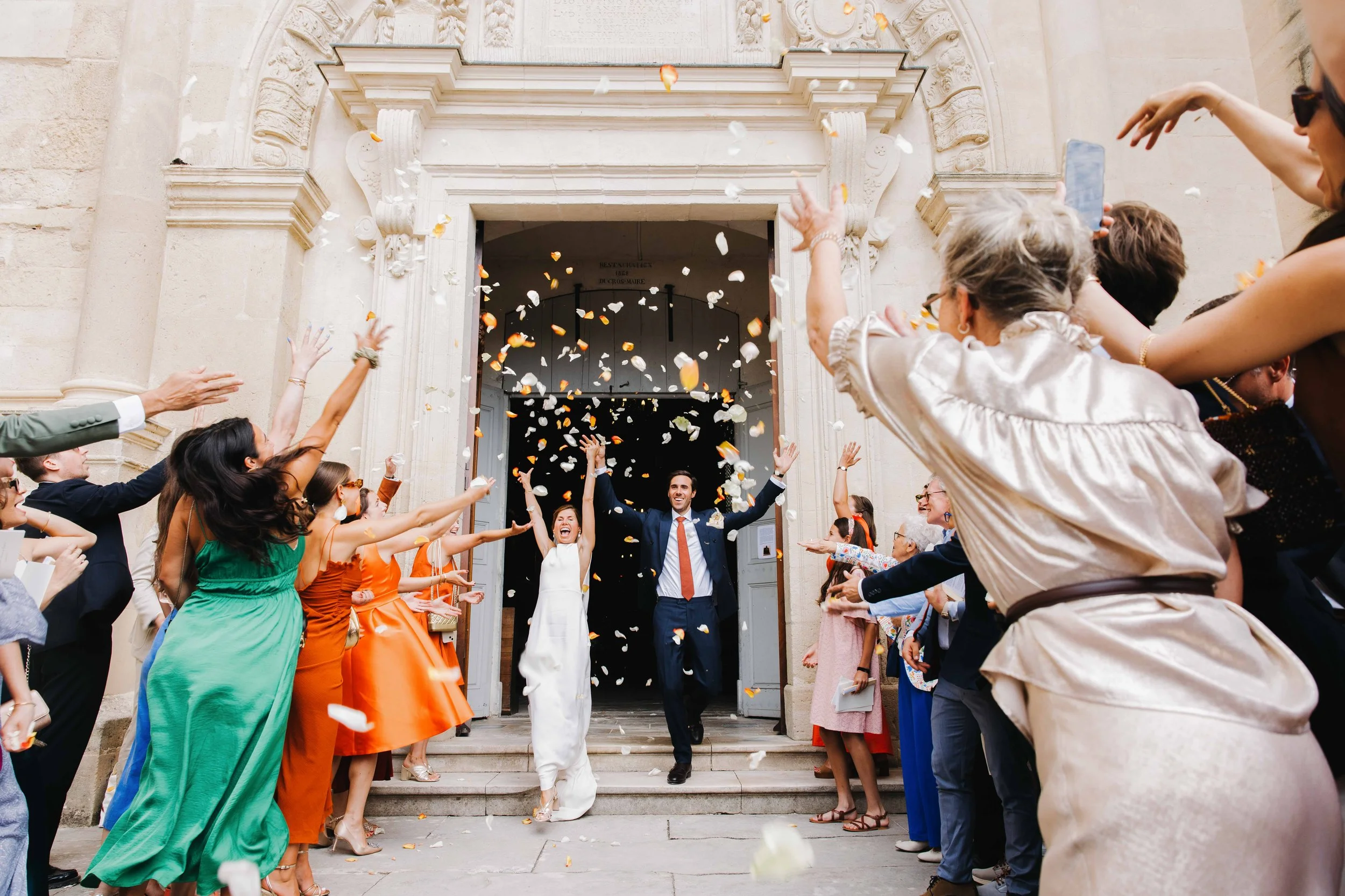 Un couple de mariés faisant leur sortie de l'église sous un arc  avec des invités lançant des pétales de fleurs en l'air. Domaine du bijoutier à grignan, drome provençal, coucher de soleil, session couple, mariage religieux, cérémonie religieuse 