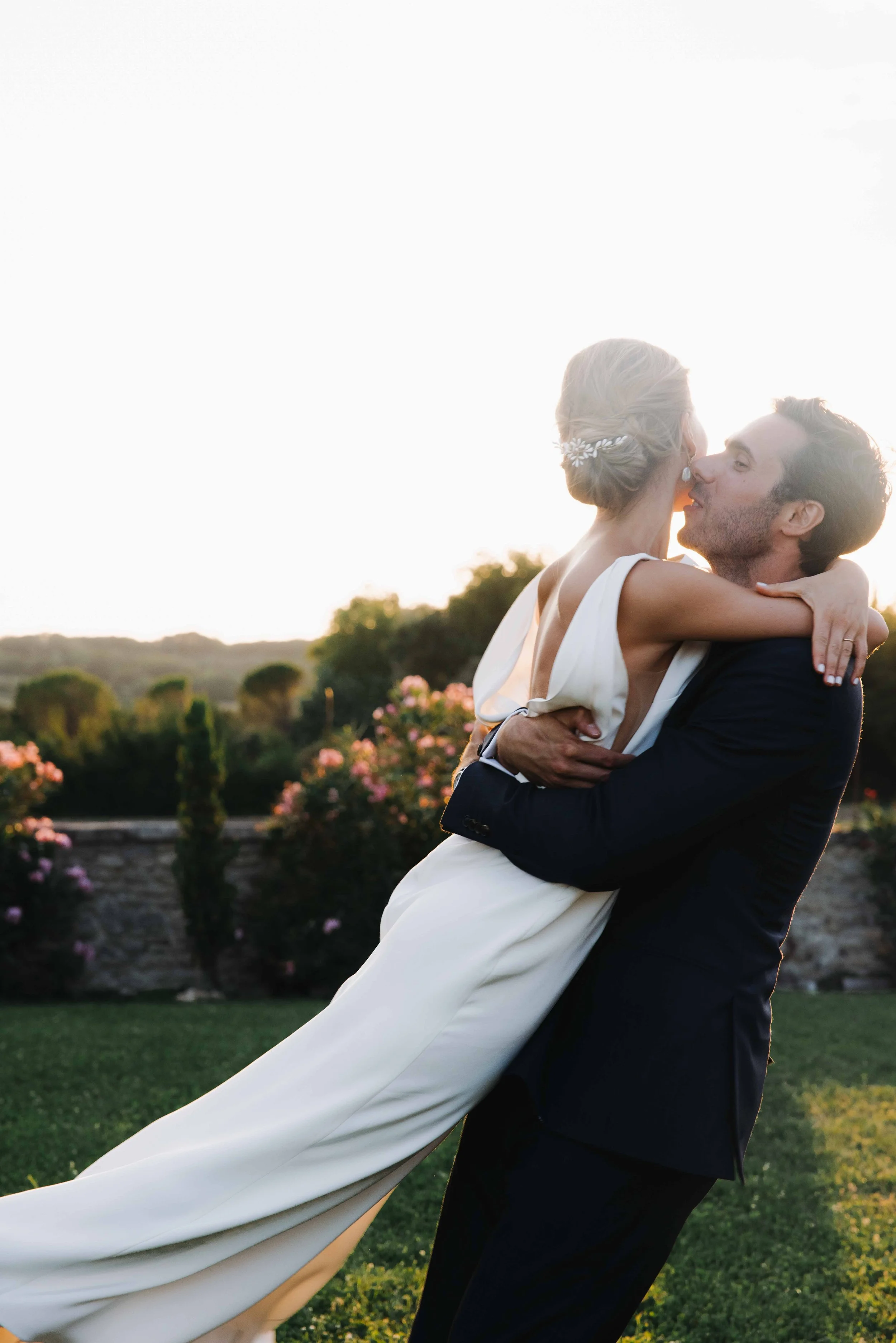 Un couple en costume de mariage, elle en robe blanche, lui en costume noir, partage un moment romantique en plein air au coucher du soleil. Domaine du bijoutier à grignan, drome provençal, coucher de soleil, session couple, mariage religieux 