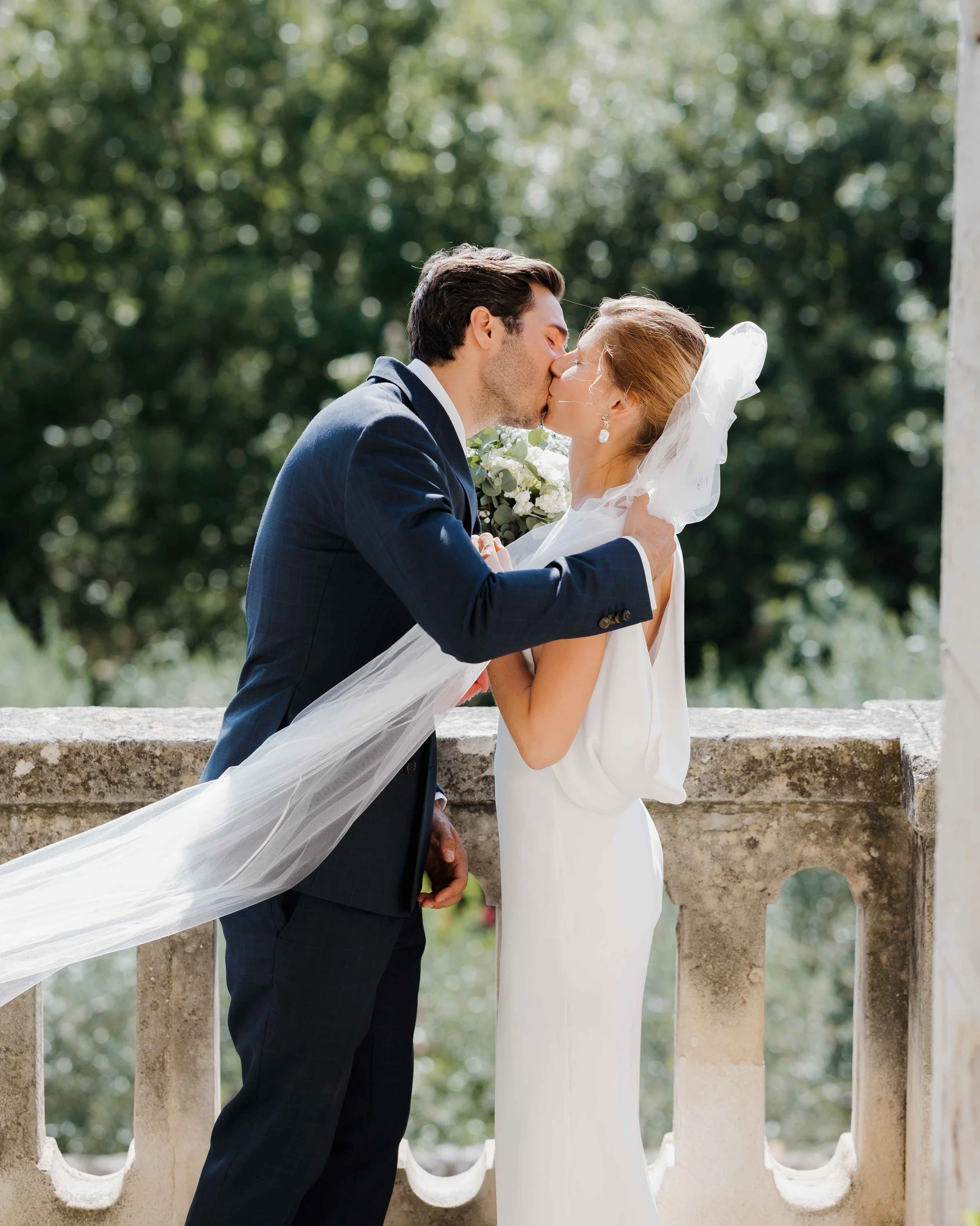 Un couple en mariage s'embrassant tendrement en extérieur, avec la femme portant une robe blanche et un voile, et le homme en costume sombre, devant une balustrade en pierre et un arrière-plan feuillu. Domaine du bijoutier à grignan, drome provençal 
