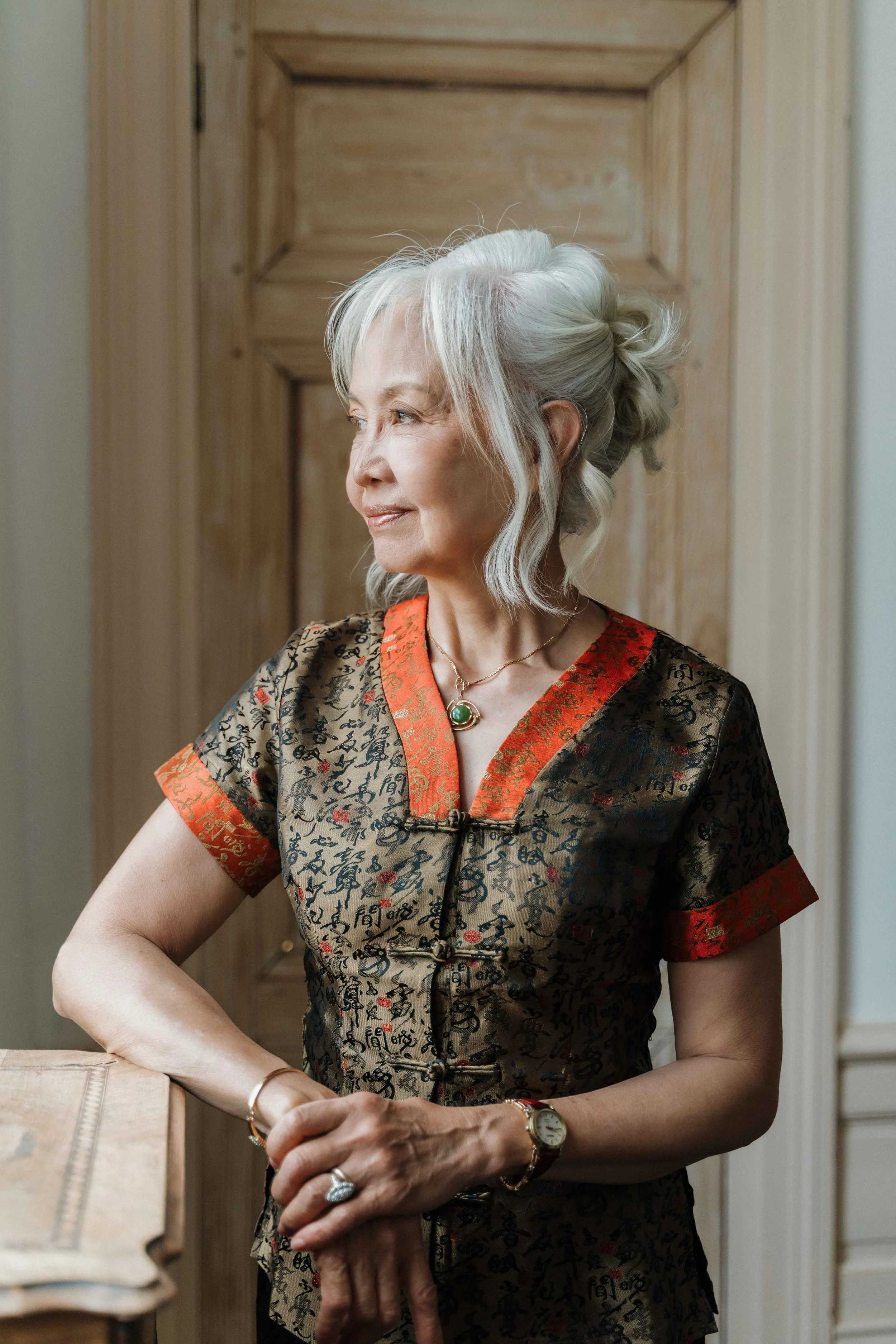 Une femme âgée aux cheveux gris portant une robe chinoise avec des motifs, regardant par la fenêtre, avec une montre en bracelet et un collier avec une pierre verte. Domaine de tourieux à Savigny dans le beaujolais à Lyon. Mariage cérémonie laïque