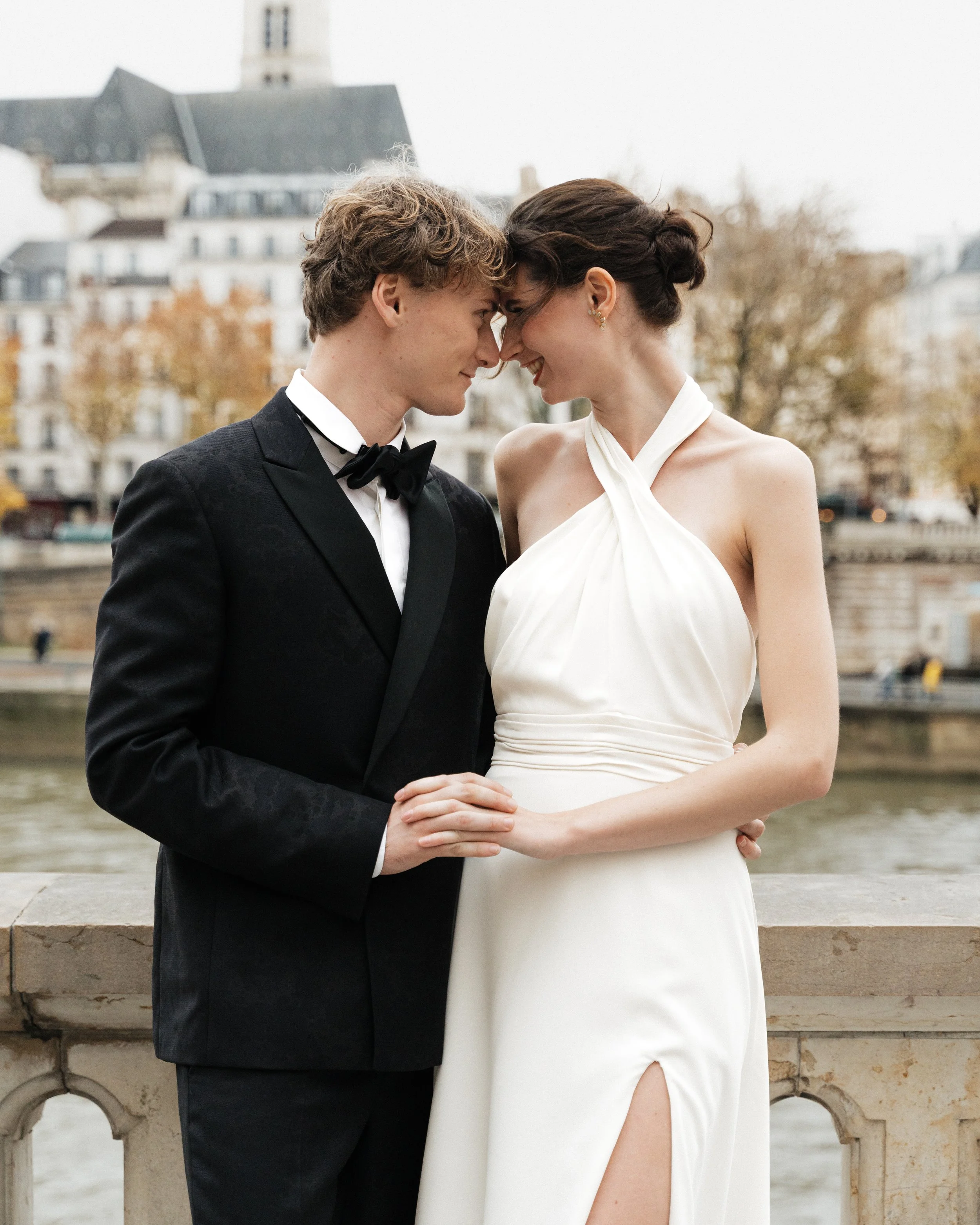 Un couple habillé en costume de mariage, se tenant très près, le front touchant, souriant, en Paris, avec un bâtiment historique en arrière-plan.