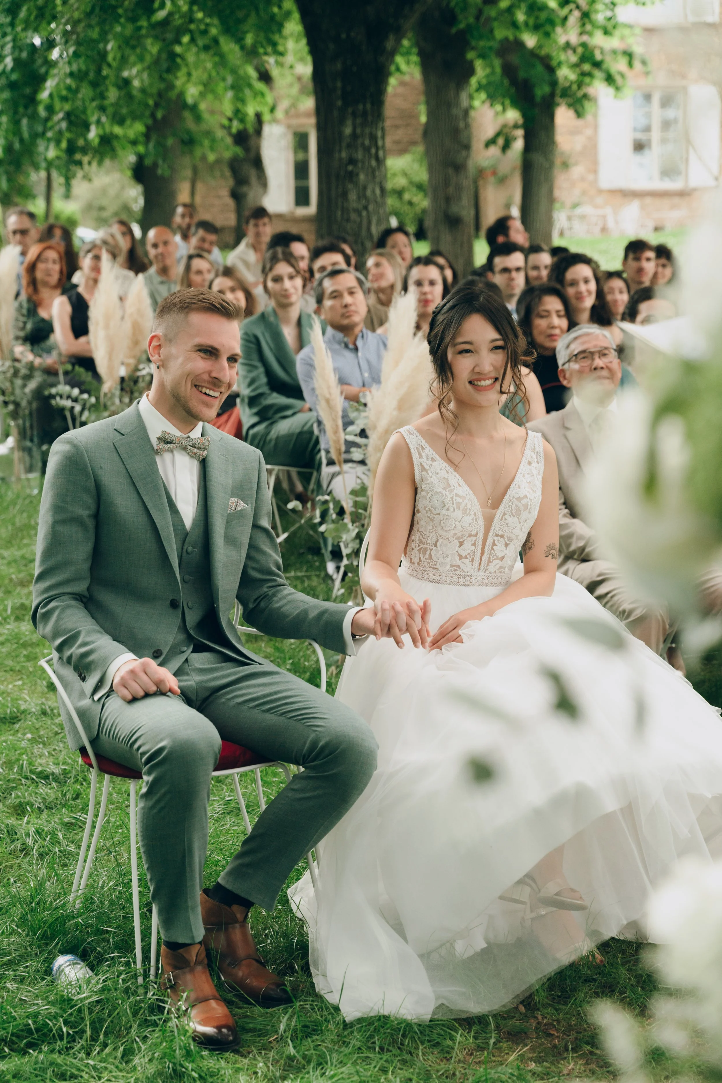 Un couple de mariés assis lors d'un mariage en plein air, entouré d'invités, sous des arbres avec un bâtiment en arrière-plan. Domaine de tourieux à Savigny dans le beaujolais à Lyon. Mariage cérémonie laïque, photographe Lyon haut de gamme, duo 