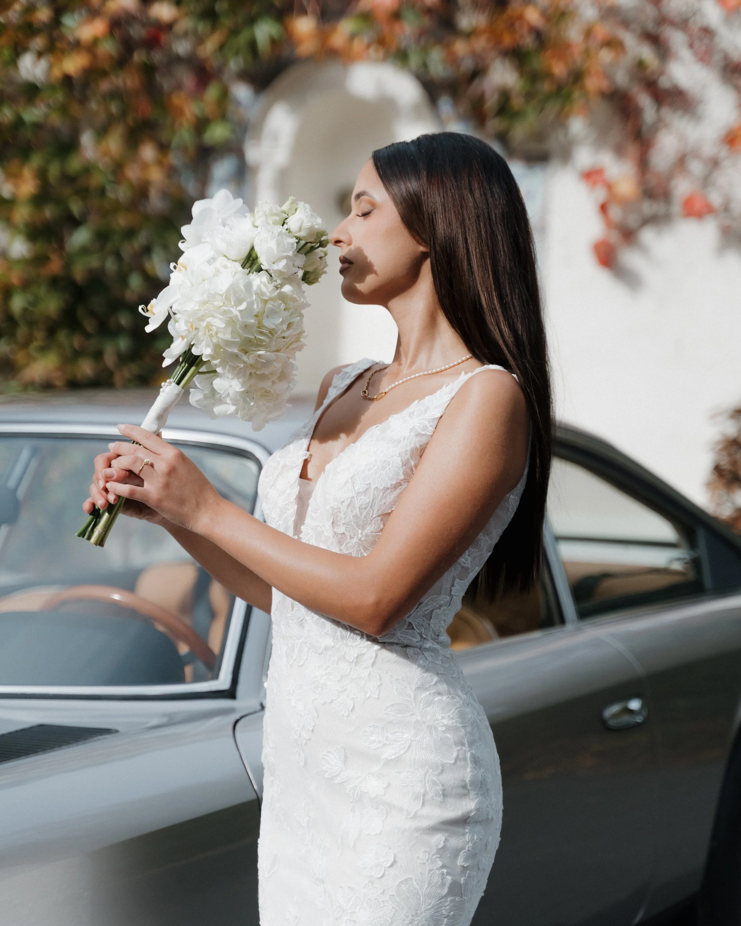 Femme en robe de mariée blanche tenant un bouquet de fleurs blanches devant une voiture classique, en extérieur.  session femme mariées et robes haut de gamme, brindos, hôtel et lac,  photographe mariage bordeaux et Lyon et destination wedding