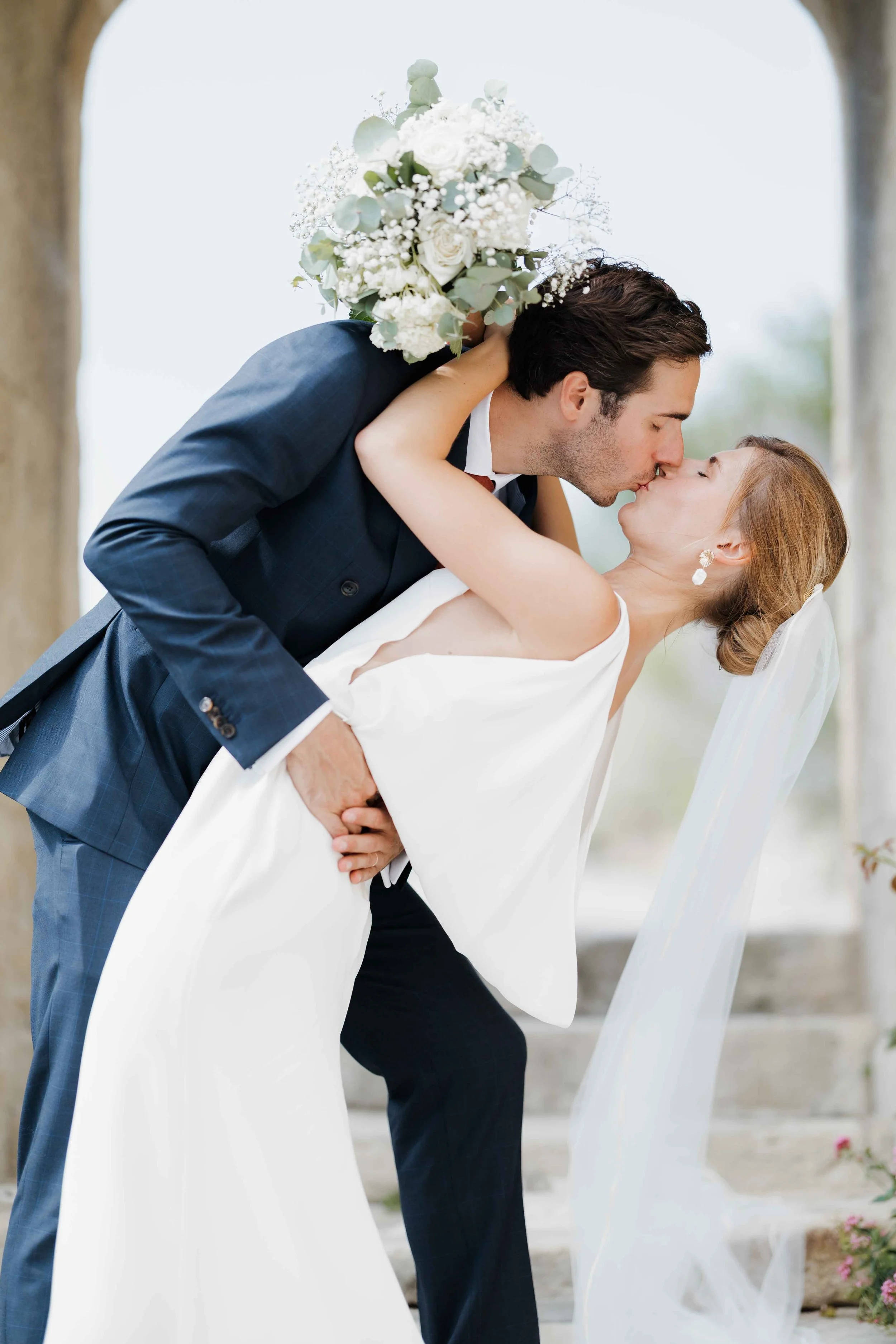 Un couple de mariés s'embrassant lors de leur mariage, l'homme en costume sombre et la femme en robe blanche, avec un bouquet de fleurs blanches. Domaine du bijoutier à grignan, drome provençal, coucher de soleil, session couple, mariage religieux 