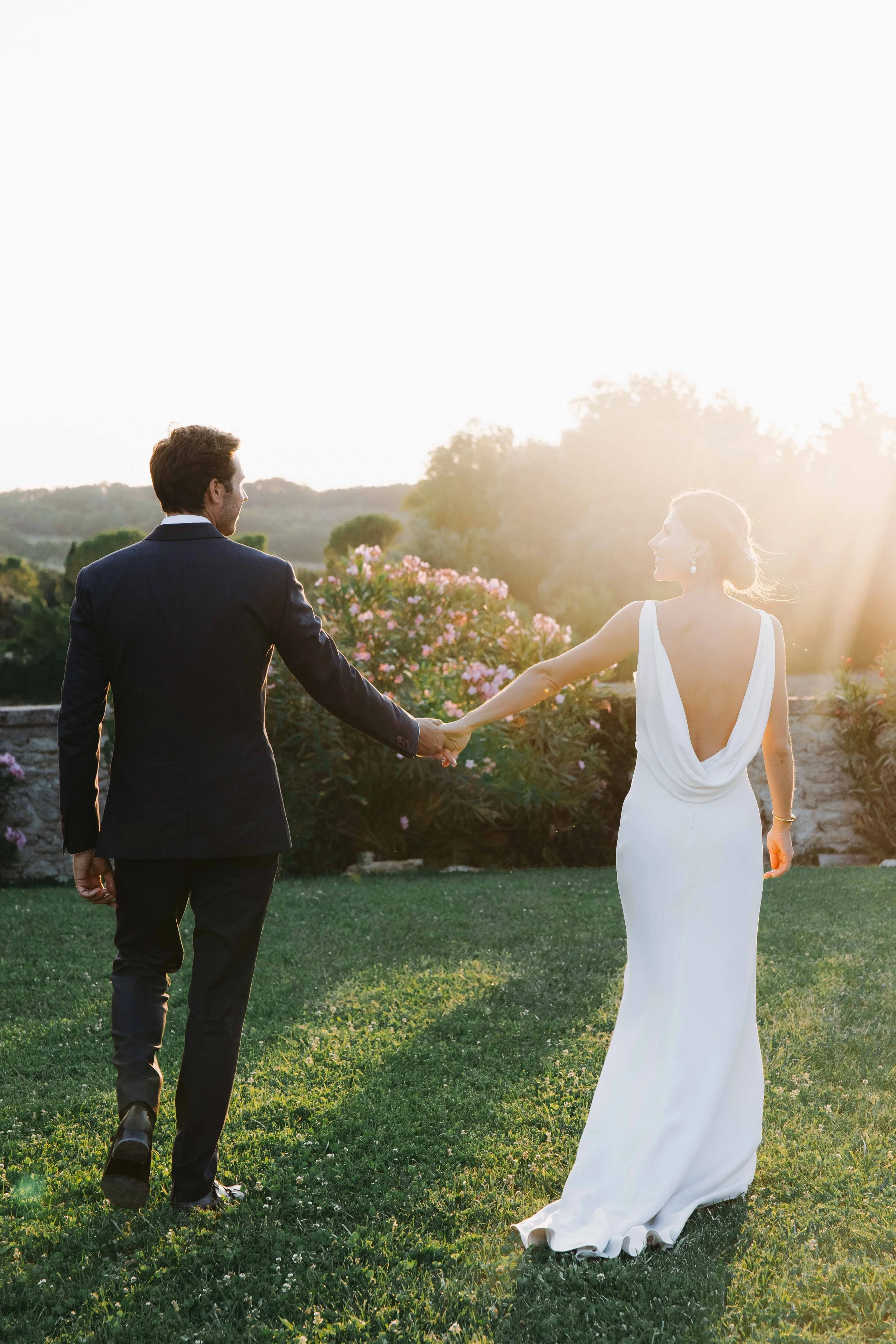 Un couple en mariage se tenant la main dans un jardin, avec le soleil couchant en arrière-plan. Domaine du bijoutier à grignan, drome provençal, coucher de soleil, session couple, mariage religieux, cérémonie religieuse 