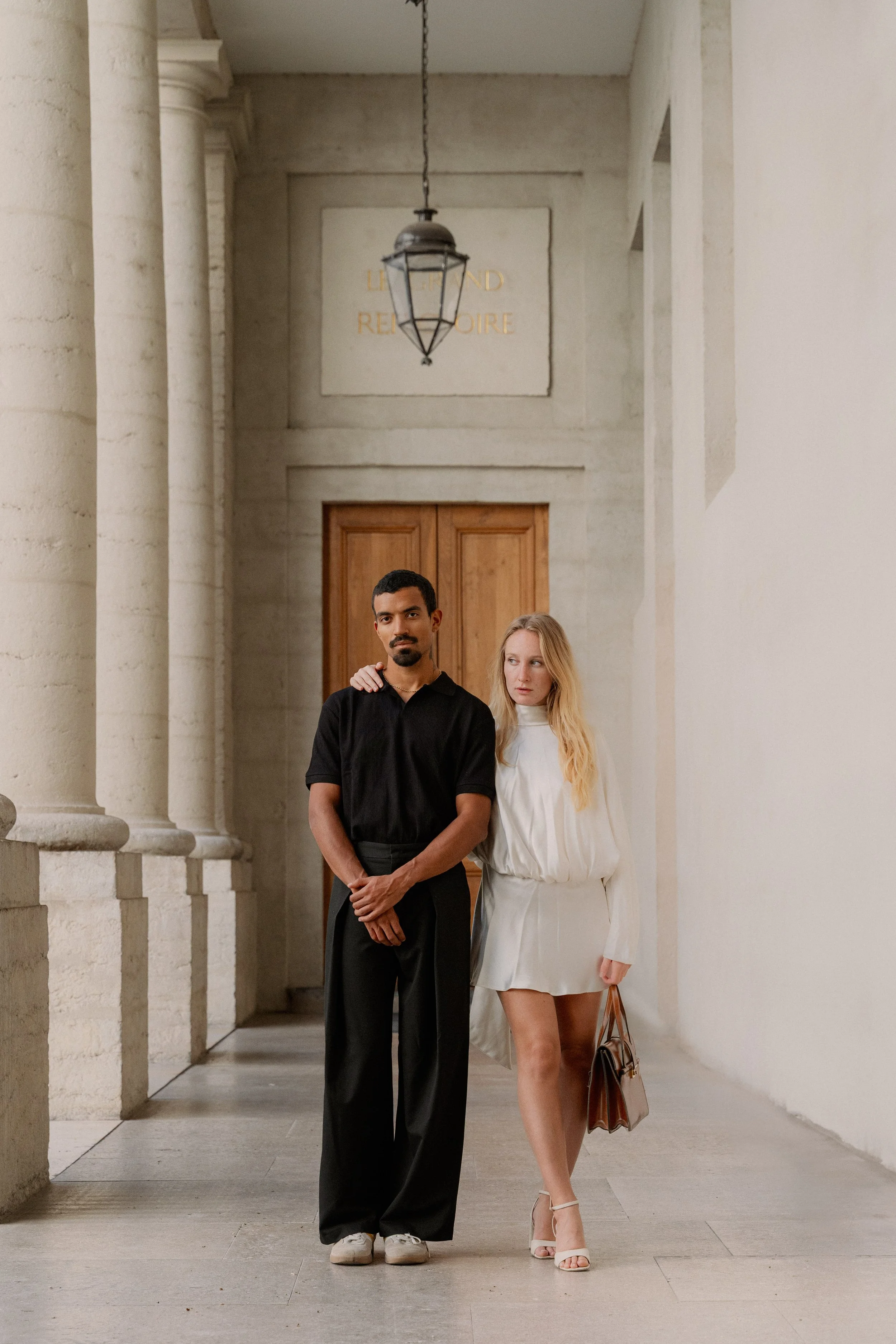 Un couple marche dans un couloir architectural, lui en noir et elle en blanc, avec une porte en bois en arrière-plan et une lampe suspendue au plafond à l'hôtel dieu à Lyon 2ème, à l'intercontinental, session engament, photographe mariage duo haut de