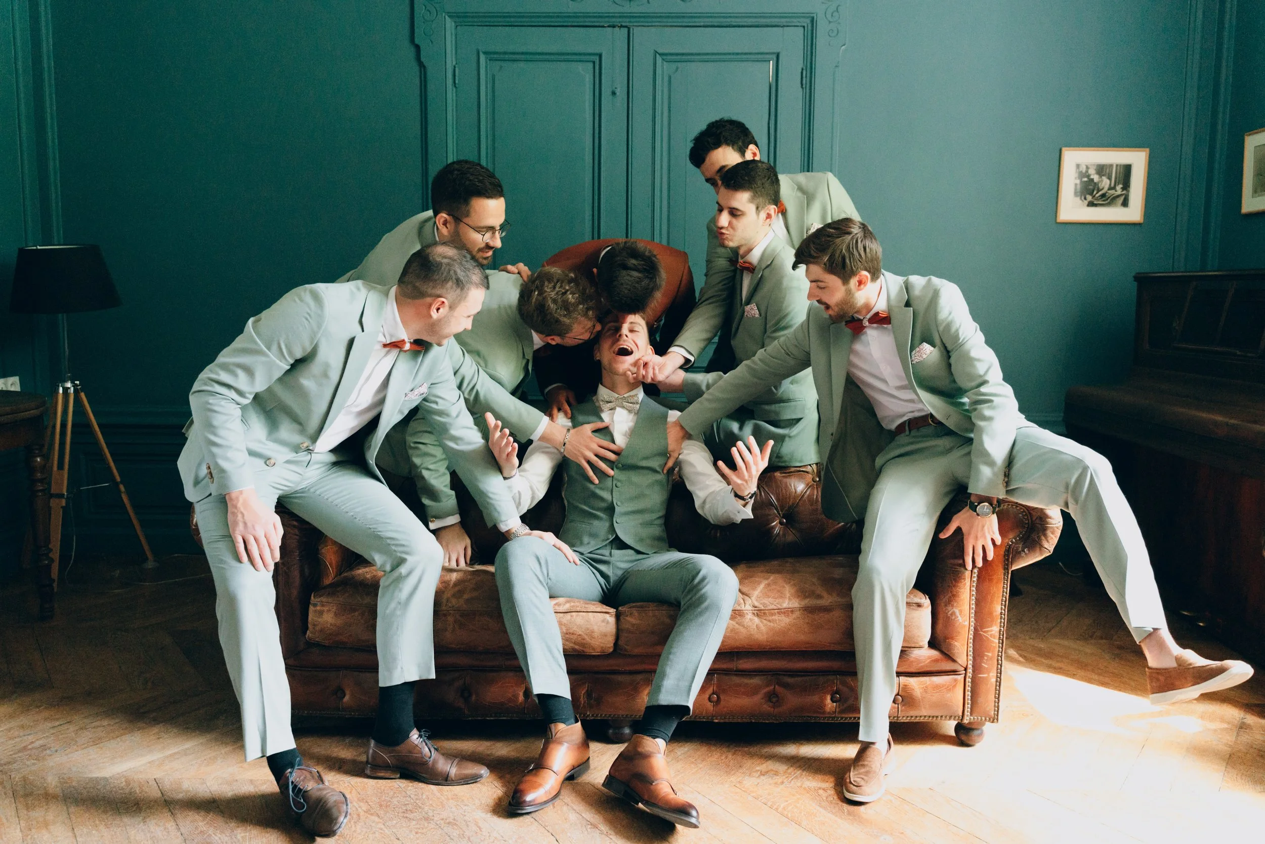 Groupe de sept jeunes hommes en costumes, entourant un autre homme assis sur un canapé, qu'ils semblent taquiner ou faire semblant de l'attaquer dans une pièce avec des murs en bois vert et deux cadres accrochés. Domaine de tourieux à Savigny 