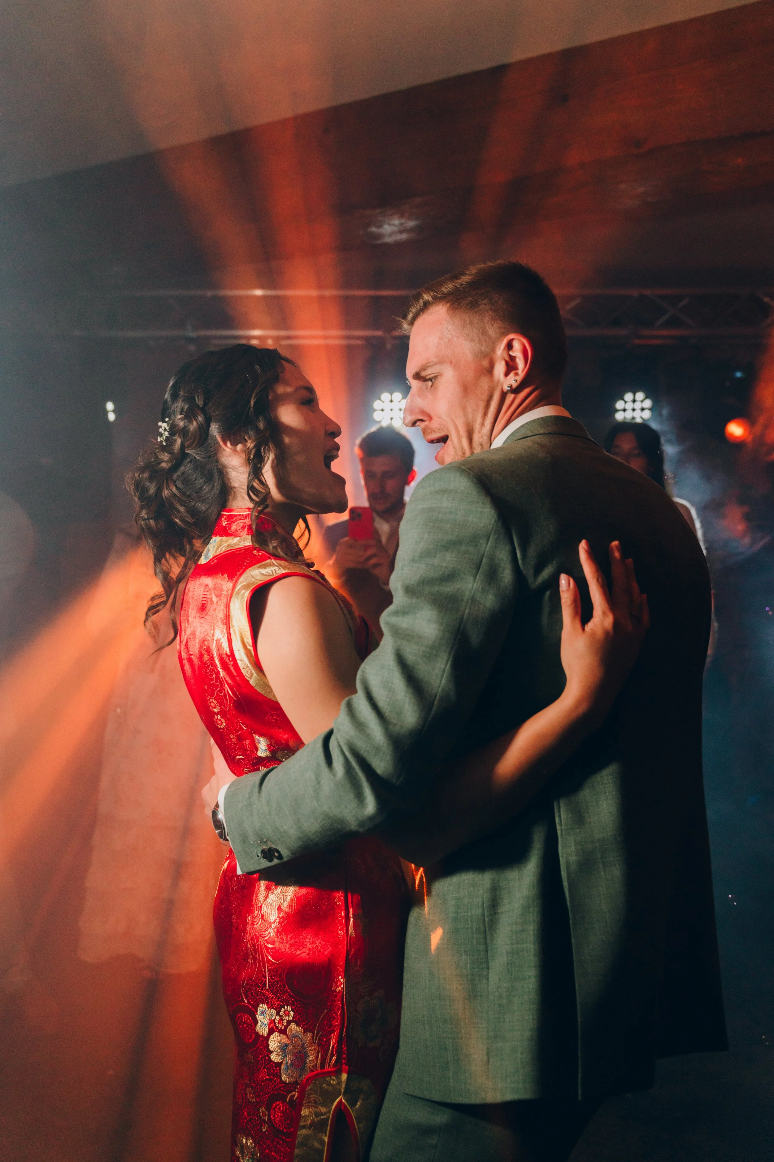 Un couple danse lors d'une fête, l'homme porte un costume gris et la femme une robe rouge traditionnelle chinoise. En arrière-plan, d'autres personnes prennent des photos en utilisant des téléphones portables, avec des lumières de scène. 
