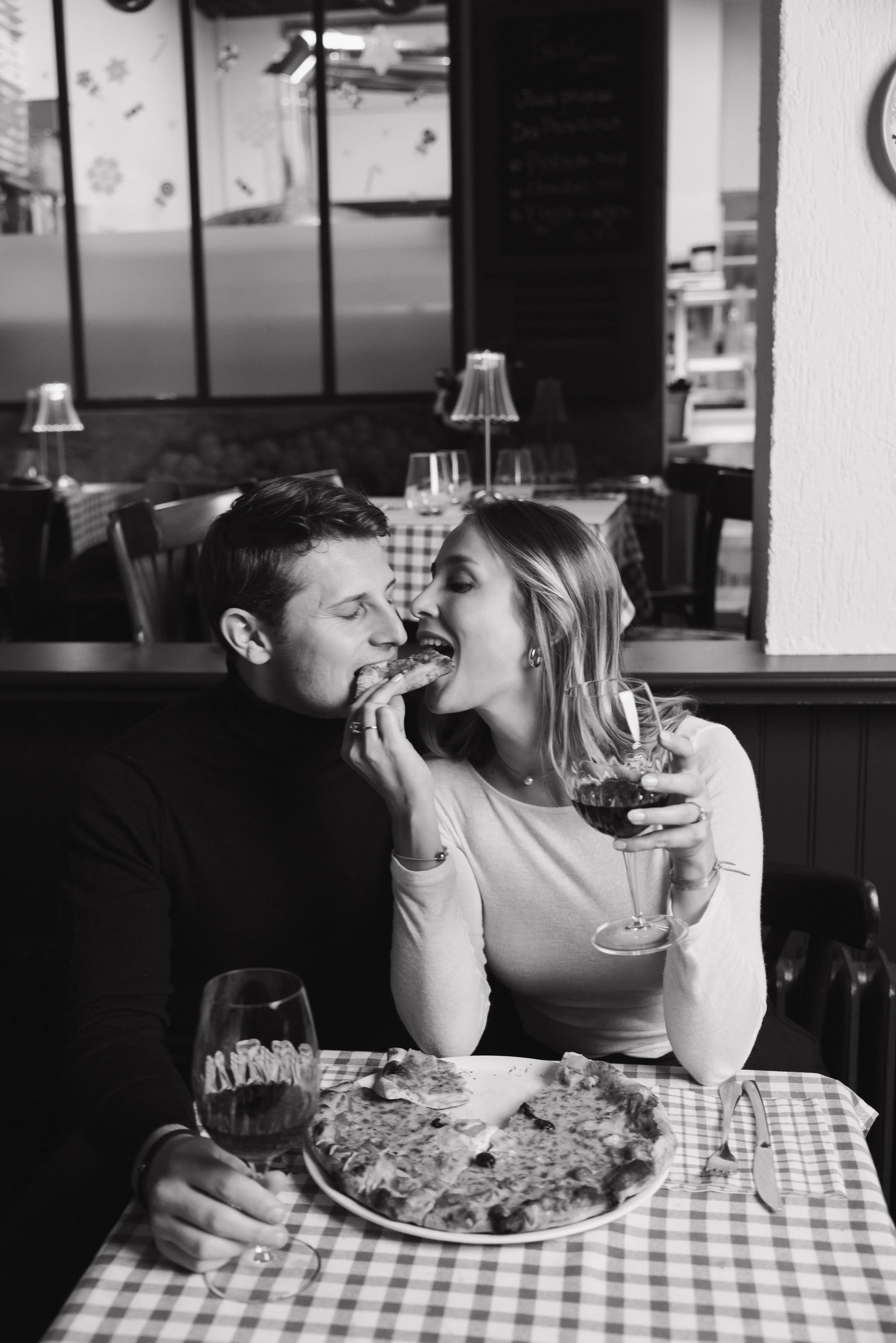 Une jeune femme et un jeune homme partagent une tranche de pizza dans un restaurant, avec une pizza et deux verres de vin rouge sur la table. session engagement couple Lyon 6eme, brotteaux, photographe mariage bordeaux et Lyon et destination wedding