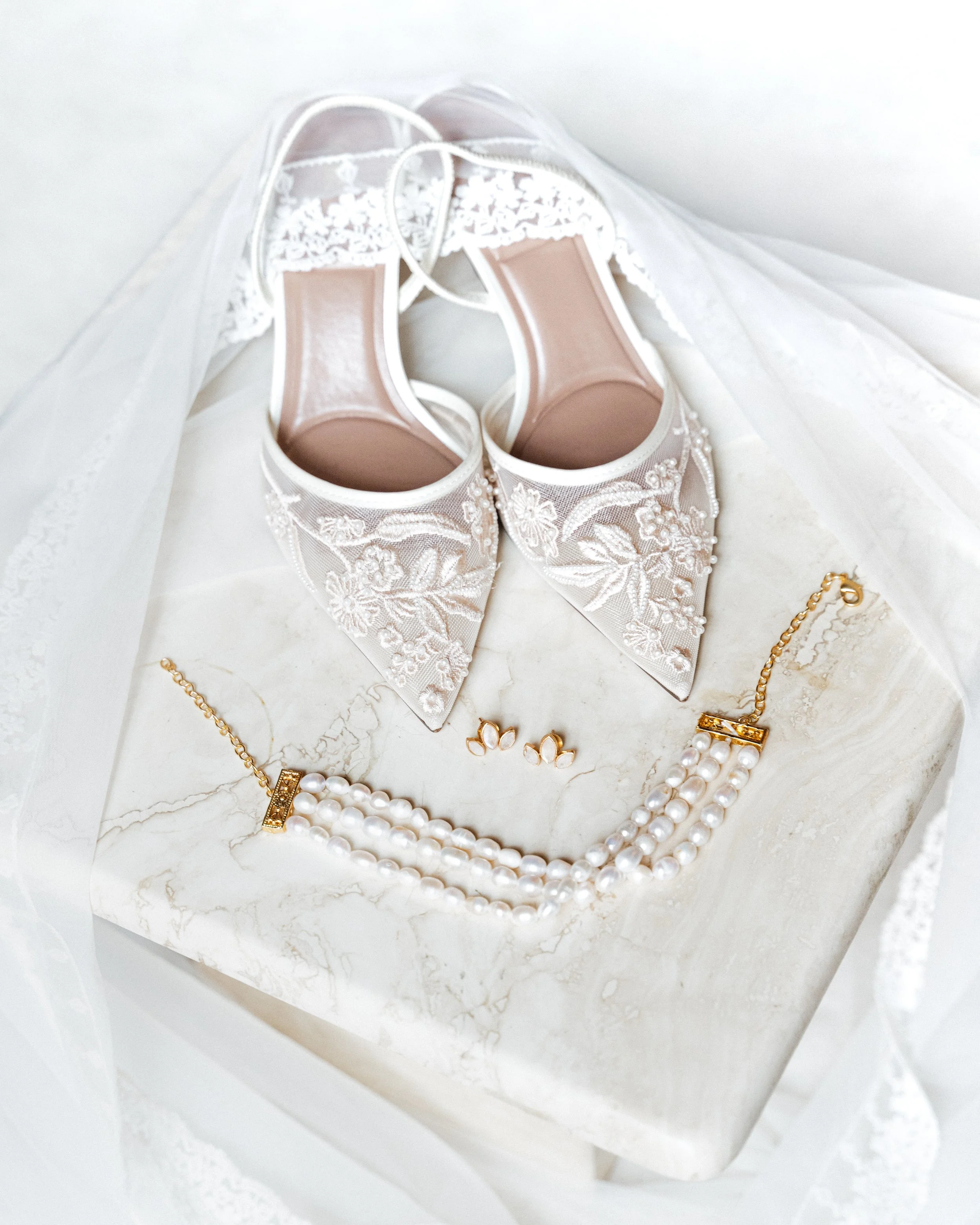 Chaussures de mariage en dentelle, collier de perles, boucles d'oreilles en perles, et une voilette blanche posés sur une surface en marbre.
