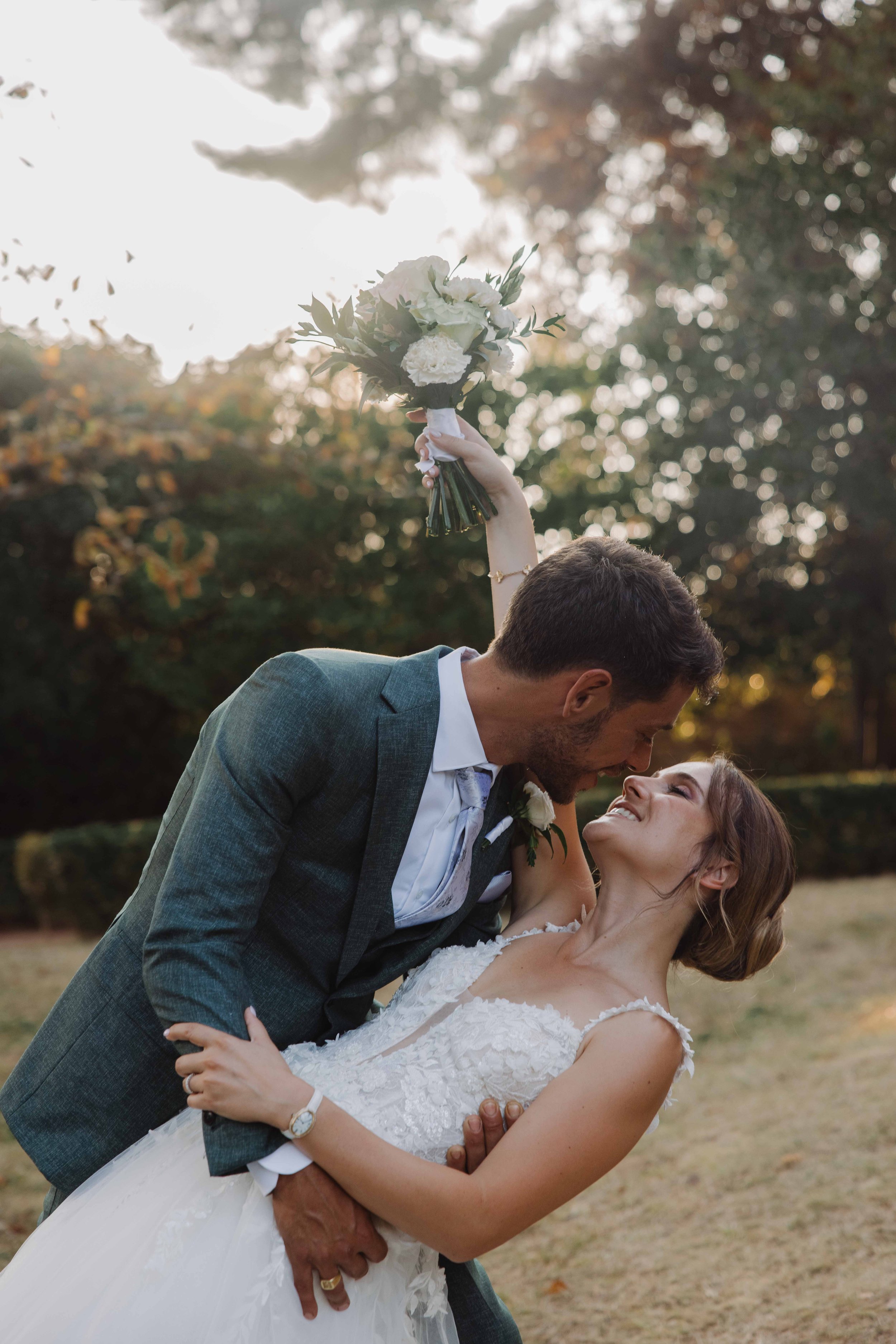 Un couple de mariés en séance photo en plein air, l'homme portant un costume gris foncé et la femme une robe de mariée blanche, en train de sourire et de s'embrasser.  Mariage chateau de champ renard, Blacé, beaujolais, Lyon, destination wedding