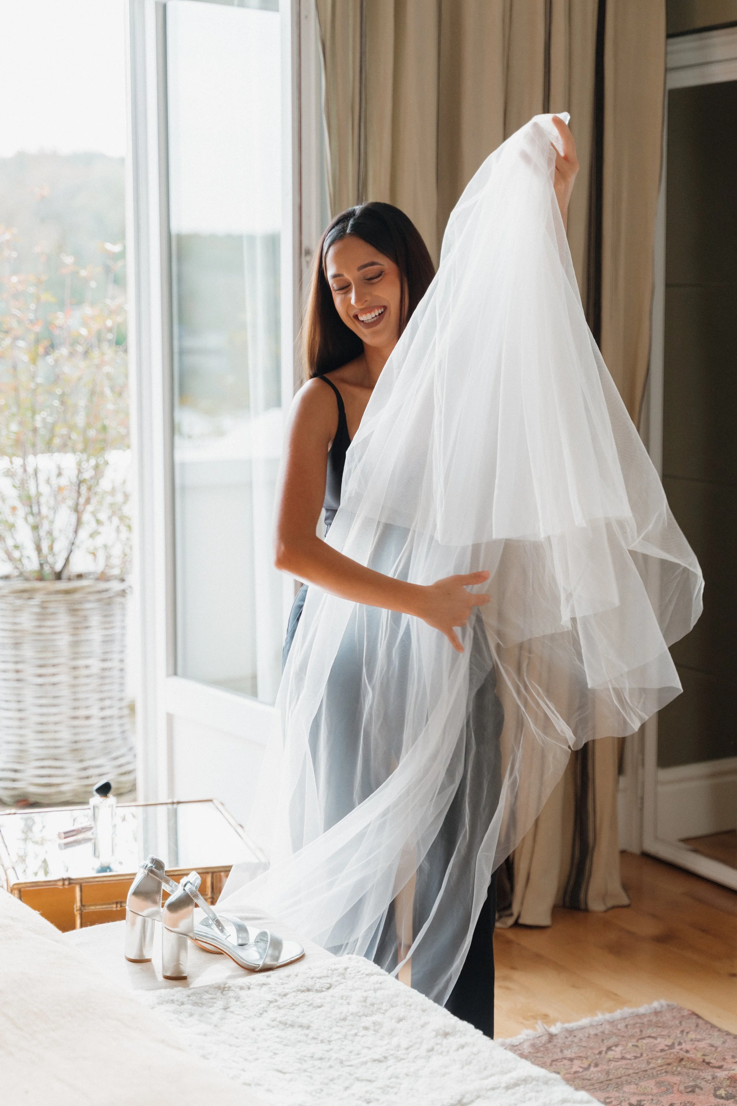 Une femme souriante essaie une robe de mariée en tulle dans une pièce bien éclairée par la lumière naturelle depuis un balcon.  session femme mariées et robes haut de gamme, brindos, hôtel et lac,  photographe mariage bordeaux et Lyon et destination