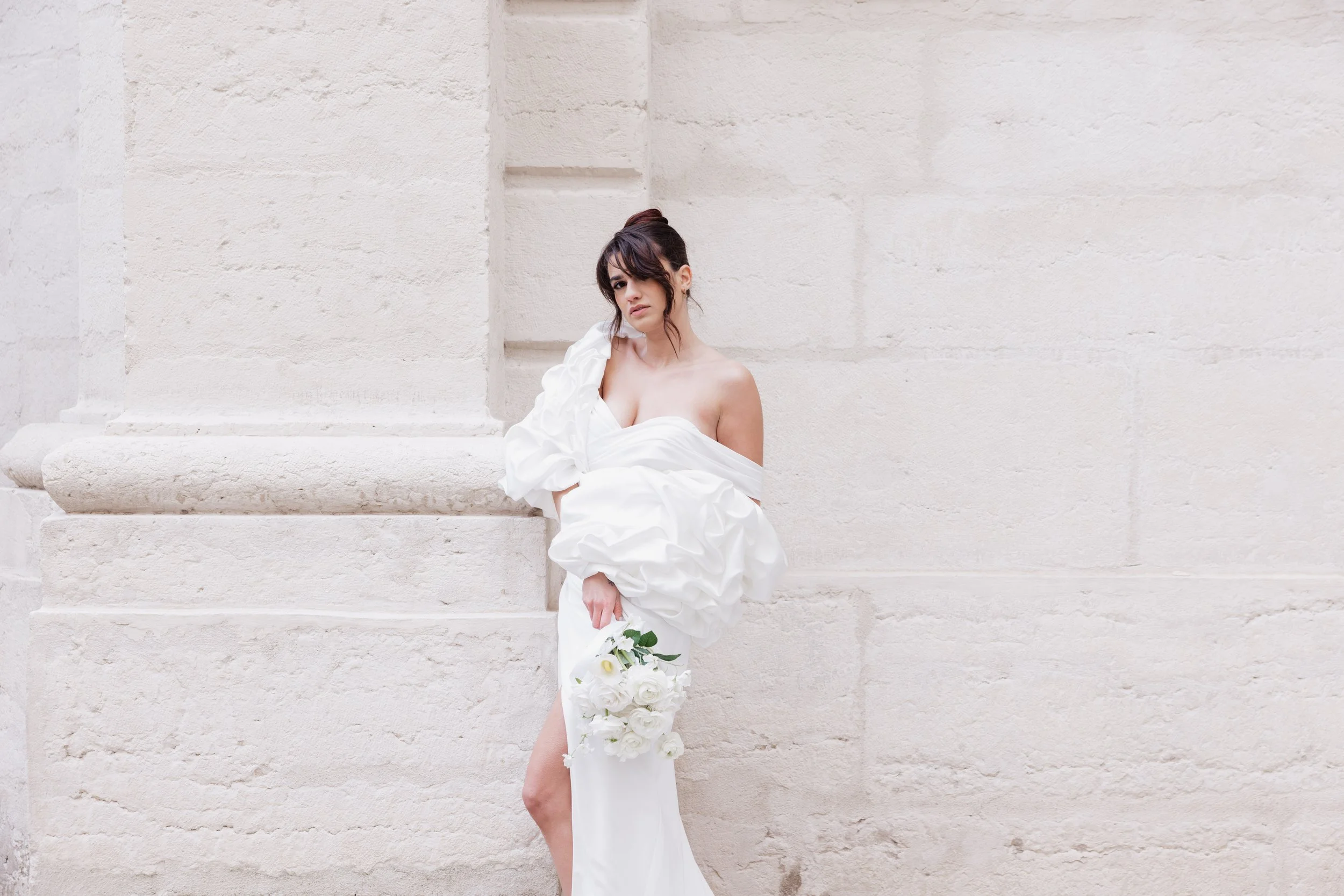 Femme en robe de mariage blanche posant contre un mur de pierre, tenant un bouquet de fleurs blanc à l'hôtel dieu à Lyon 2ème, à l'intercontinental, session engament, photographe mariage duo haut de gamme.