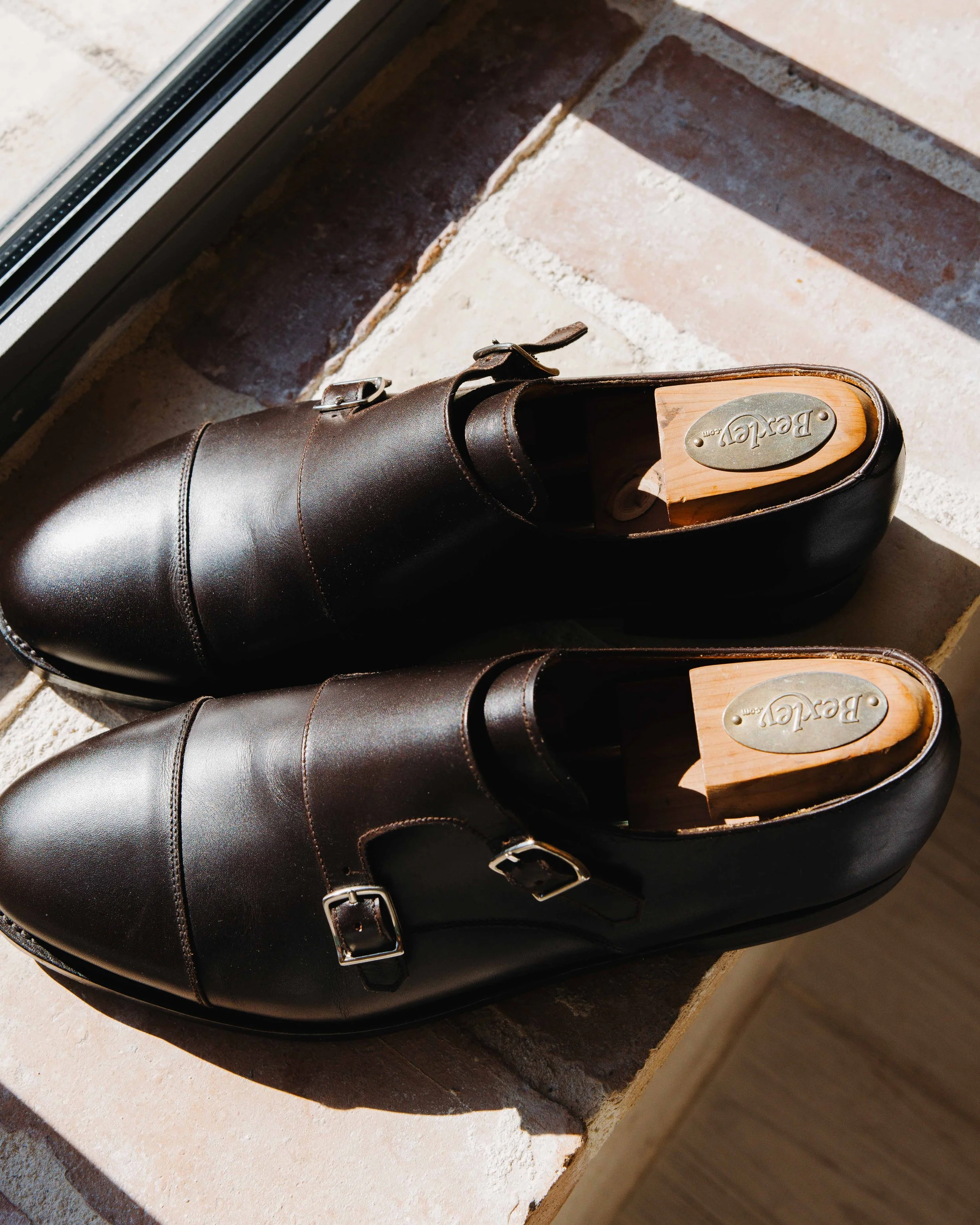 Une paire de chaussures en cuir marron foncé avec des boucles en métal, présentée sur un rebord en pierre à côté d'une fenêtre, avec des chaussures en bois à l'intérieur. Domaine du bijoutier à grignan, drome provençal, préparatifs homme 