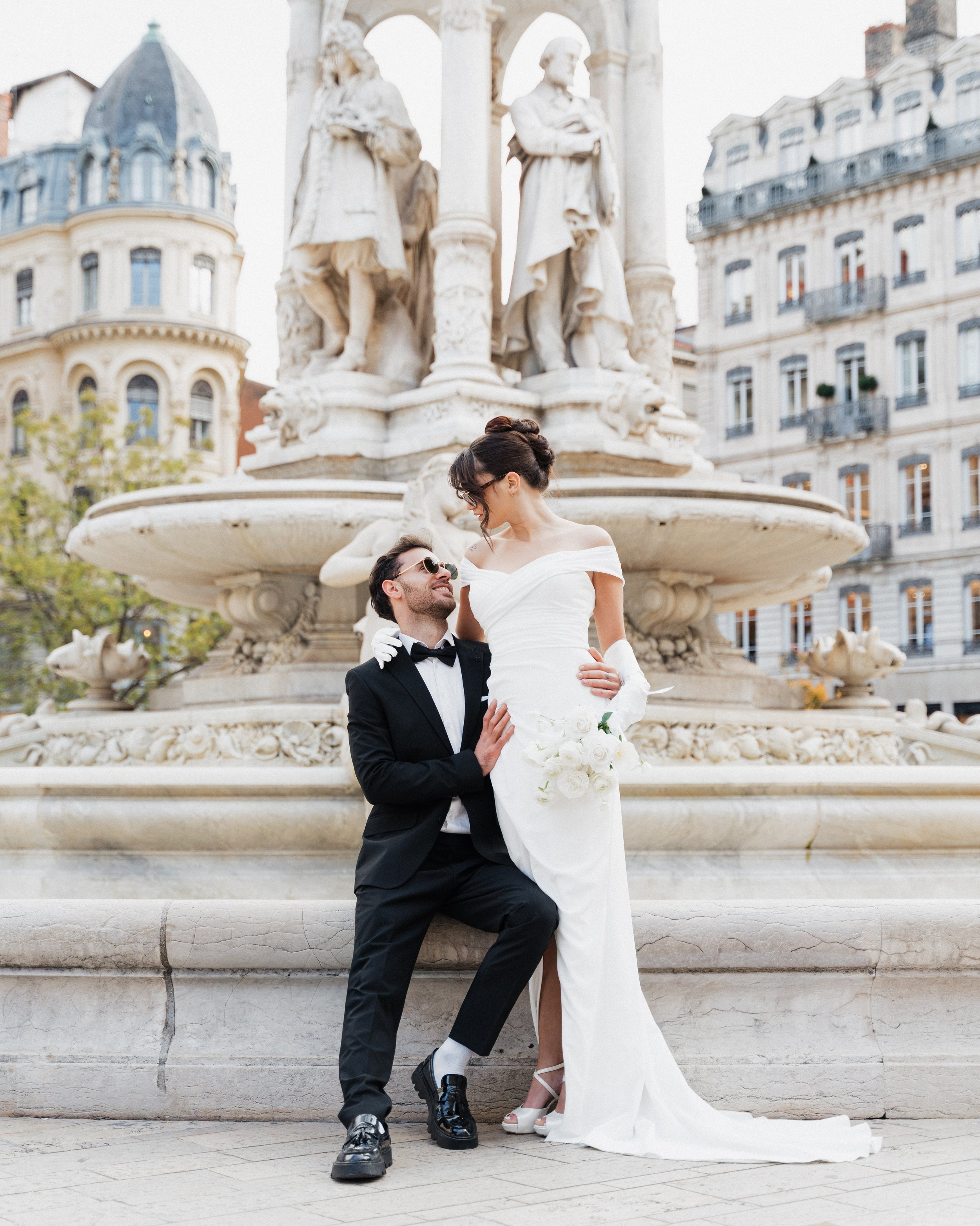 Un couple en robe de mariage et costume, posant devant une fontaine ornée de sculptures, dans une ville avec des bâtiments historiques à Lyon place des jacobins, photographe duo haut de gamme mariage Lyon et bordeaux