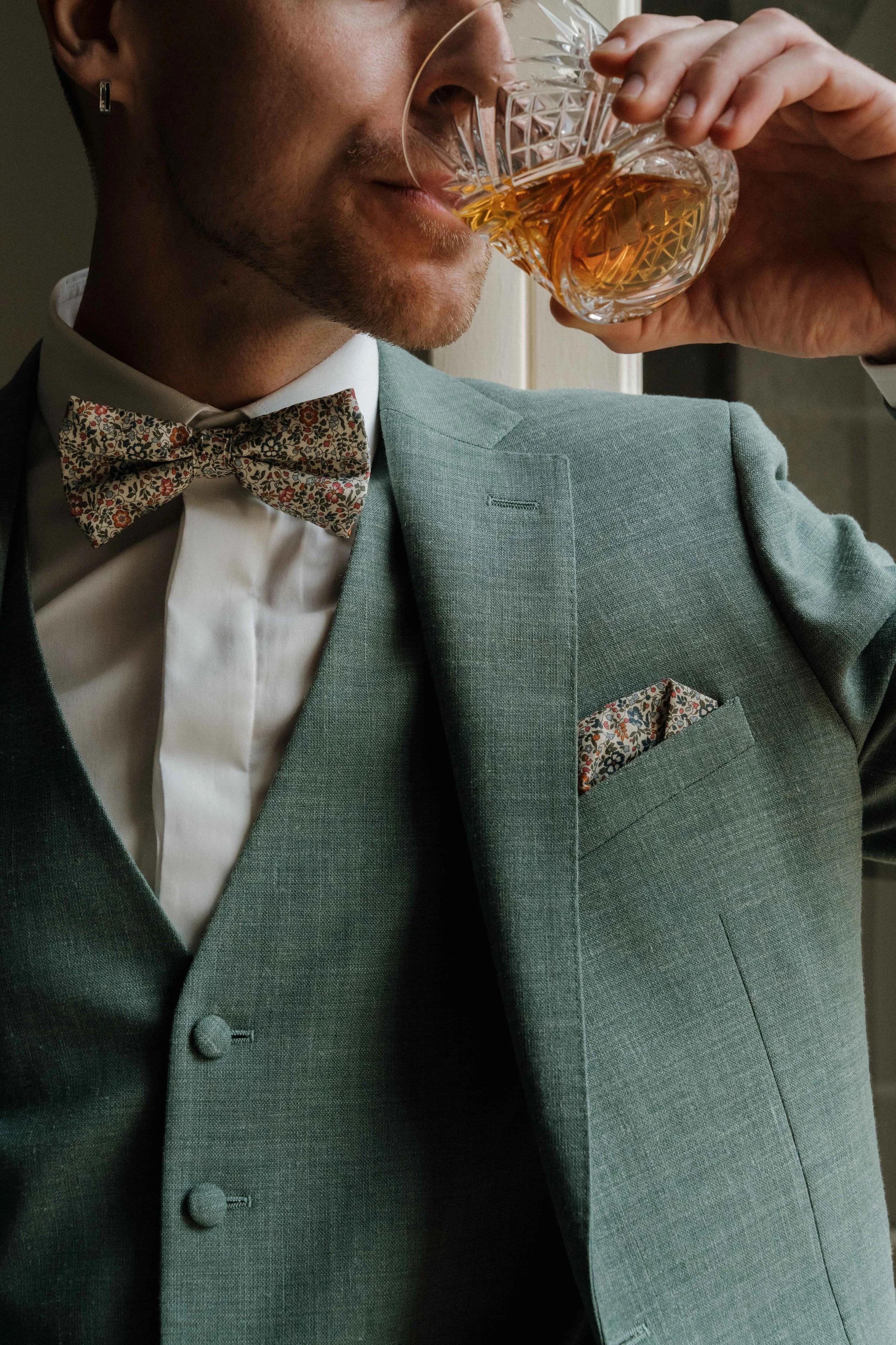 Homme en costume vert avec nœud papillon à motif floral, buvant un verre de whisky ou de bourbon. Domaine de tourieux à Savigny dans le beaujolais à Lyon. Mariage cérémonie laïque, photographe Lyon haut de gamme, duo photographe, argentique.