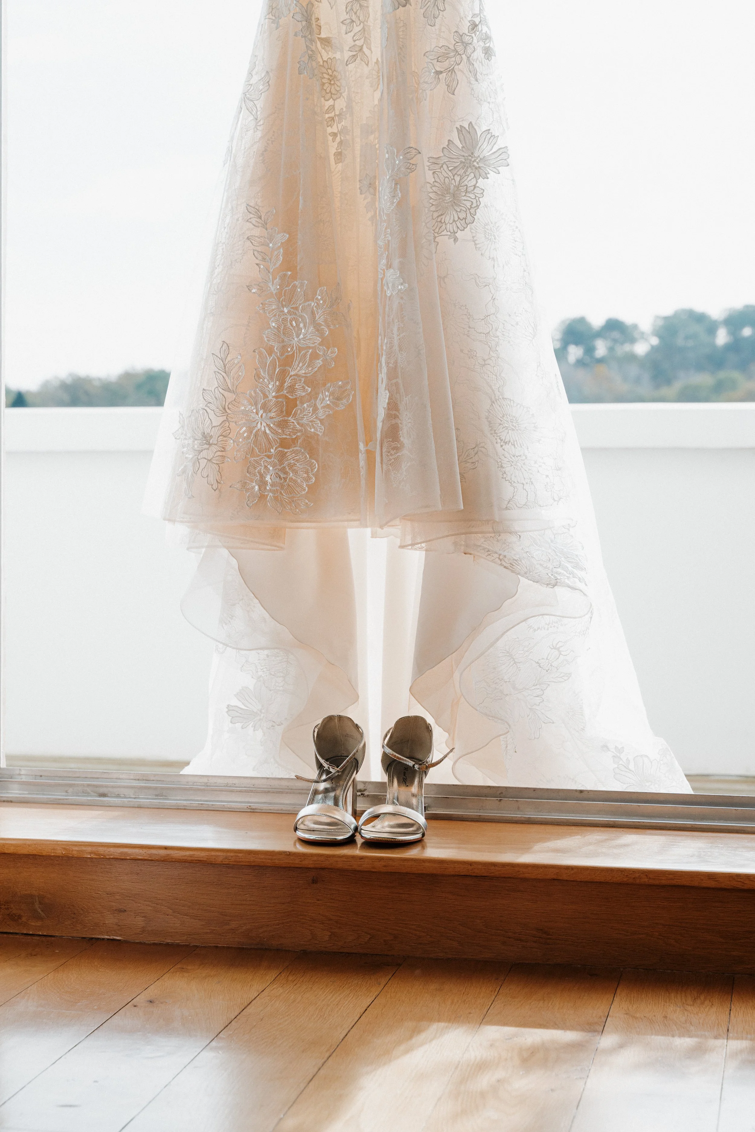 Robe de mariage suspendue devant une fenêtre, avec des chaussures à talons argentées en dessous.  session femme mariées et robes haut de gamme, brindos, hôtel et lac,  photographe mariage bordeaux et Lyon et destination wedding