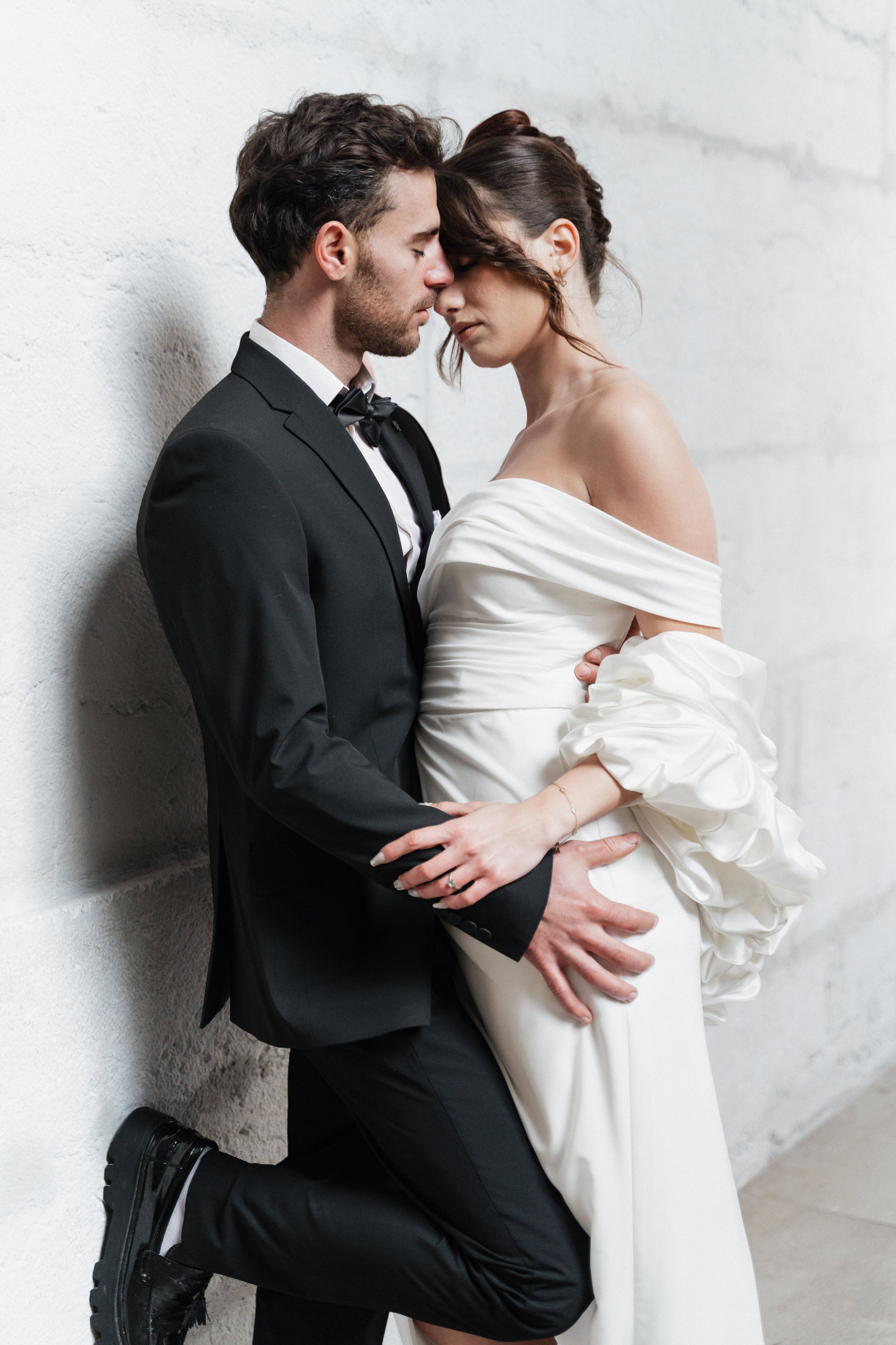 Un couple en tenue de mariage, homme en costume noir et femme en robe blanche, posant contre un mur blanc, leur tête fréquemment touchant à Lyon 2ème à l'hôtel dieu à l'intercontinental lors d'une session engagement par un duo de photographe.