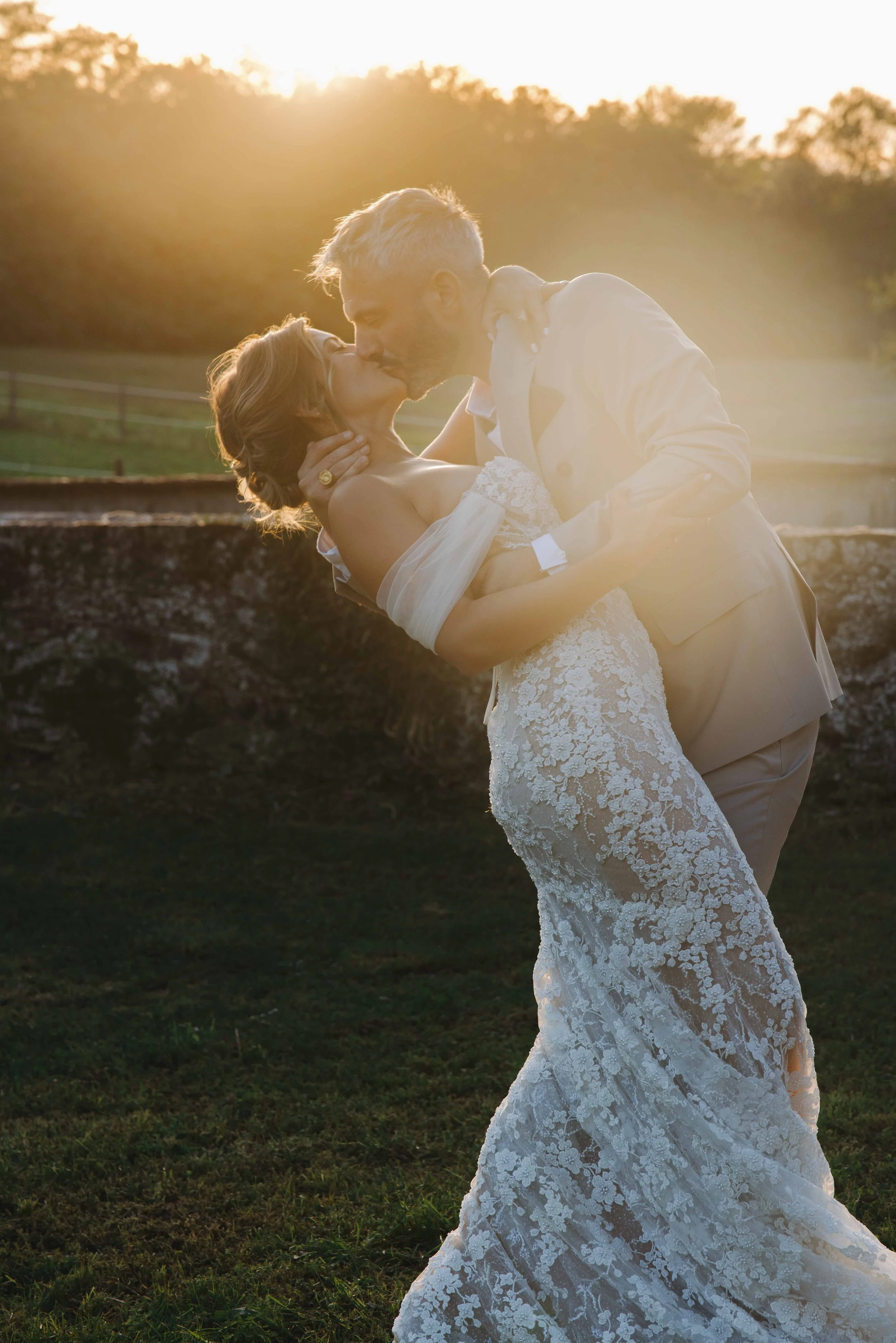 Un couple en robe de mariage partage un baiser dans un décor naturel au coucher du soleil. Mariage à la ferme du rocher en Isère, robe de mariées, coucher de soleil, photos de couples. Photographe en duo haut de gamme et destination wedding. Bordeaux