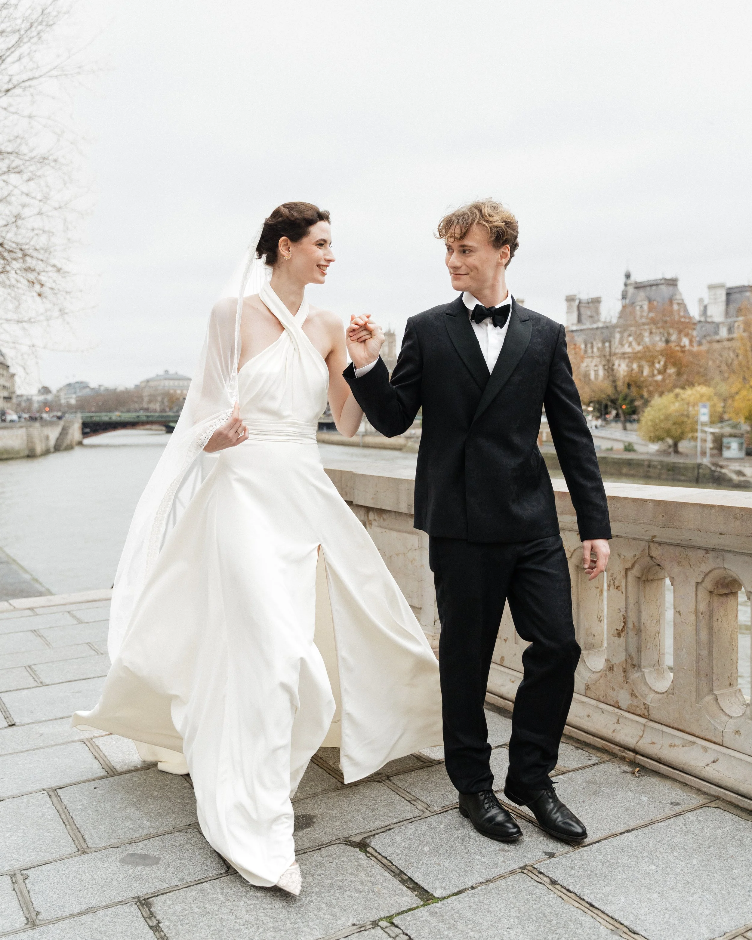 Un couple en costume de mariage marchant main dans la main sur un pont, avec la Seine et des bâtiments parisiens en arrière-plan. Session engagement paris, Louvre, mariage haut de gamme, destination wedding. Robe de mariées, madame a dit oui
