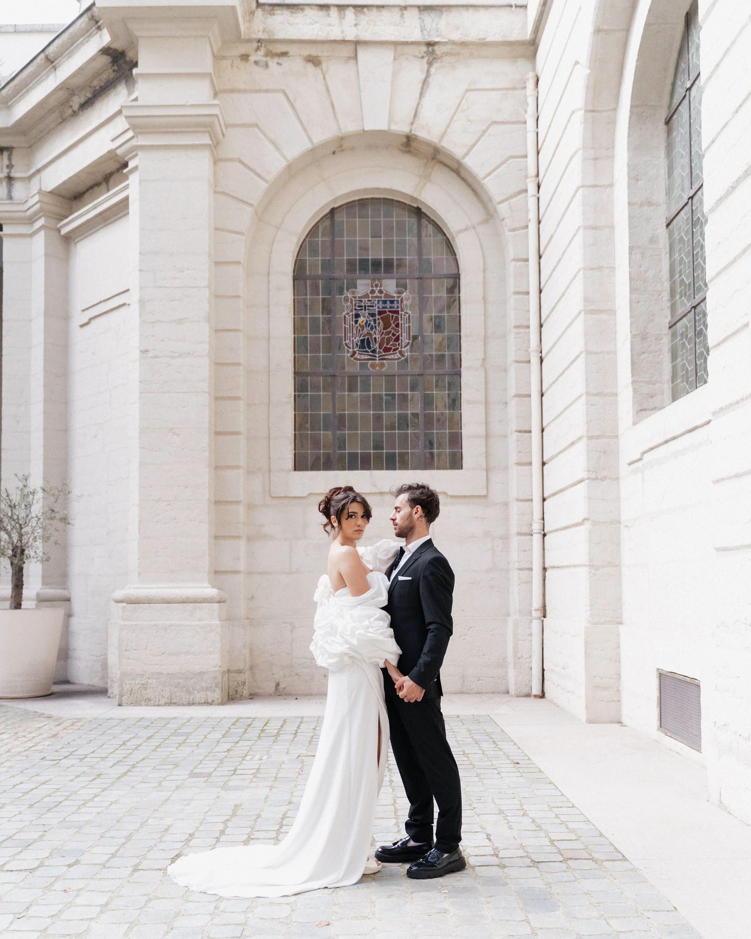 Un couple en vêtements de mariage, la femme en robe blanche et l'homme en costume noir, se tenant dans une cour pavée devant un bâtiment en pierre avec une grande fenêtre en mosaïque, lors d'une séance photo de mariage à l'hôtel dieu à Lyon 2ème.