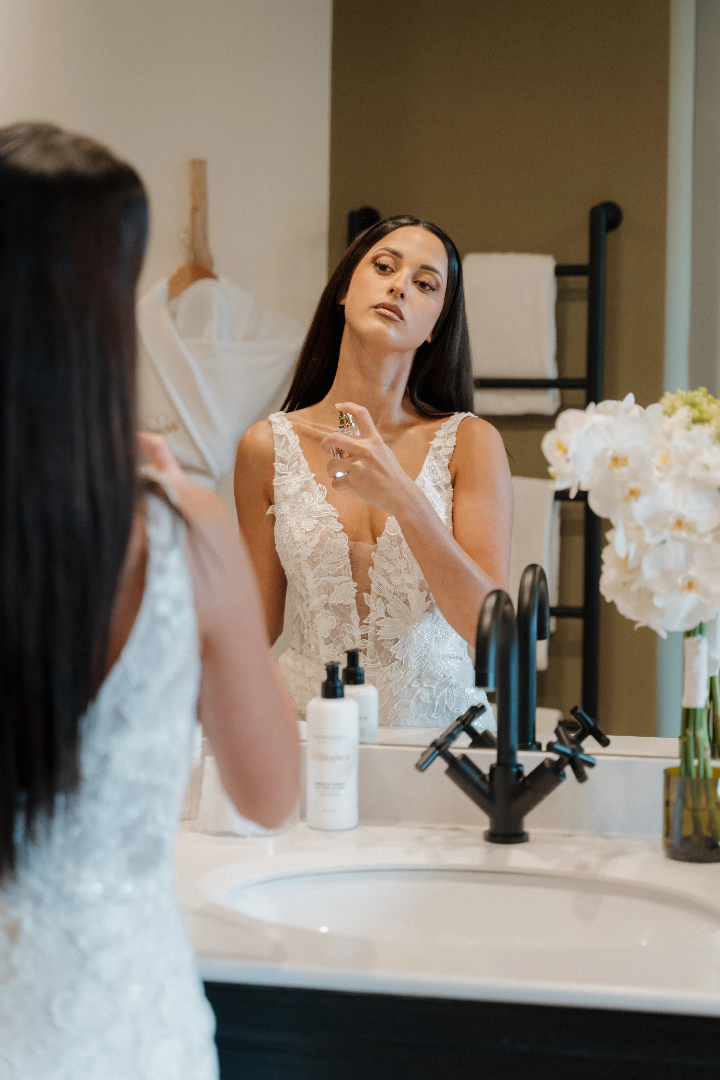 Une femme en robe de mariage regarde son reflet dans le miroir en se maquillant dans une salle de bains moderne.  session femme mariées et robes haut de gamme, brindos, hôtel et lac,  photographe mariage bordeaux et Lyon et destination wedding