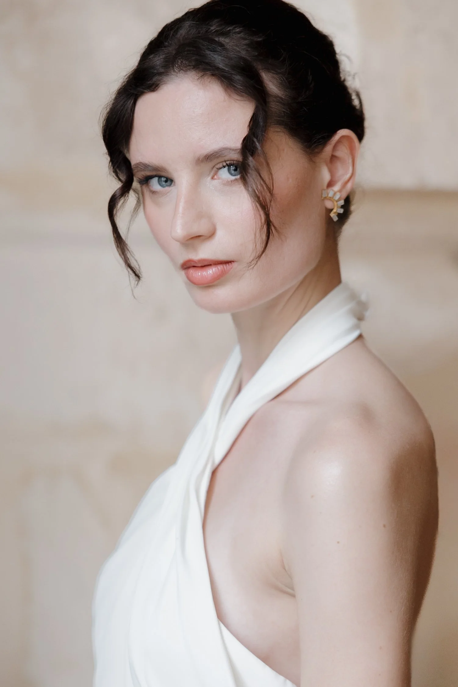 Jeune femme avec peau claire, cheveux bruns bouclés et ne passant qu'une épaulette d'une robe blanche, portant des boucles d'oreilles dorées, regardant vers la caméra dans un décor neutre. Session engagement paris, Louvre, mariage haut de gamme