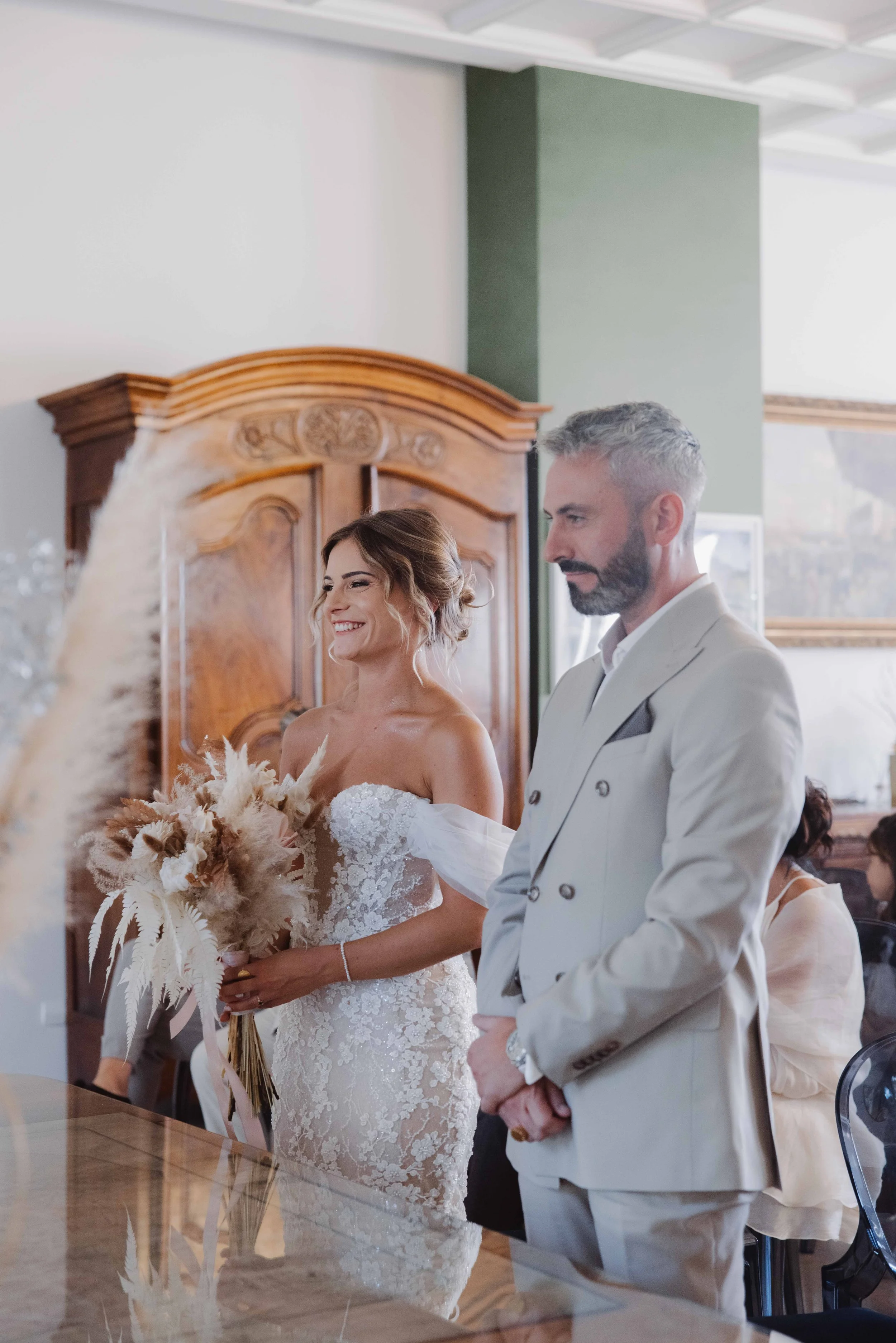 Une femme en robe de mariée souriante et un homme en costume blanc lors d'une cérémonie de mariage, dans une pièce avec mobilier en bois et portraits sur le mur.