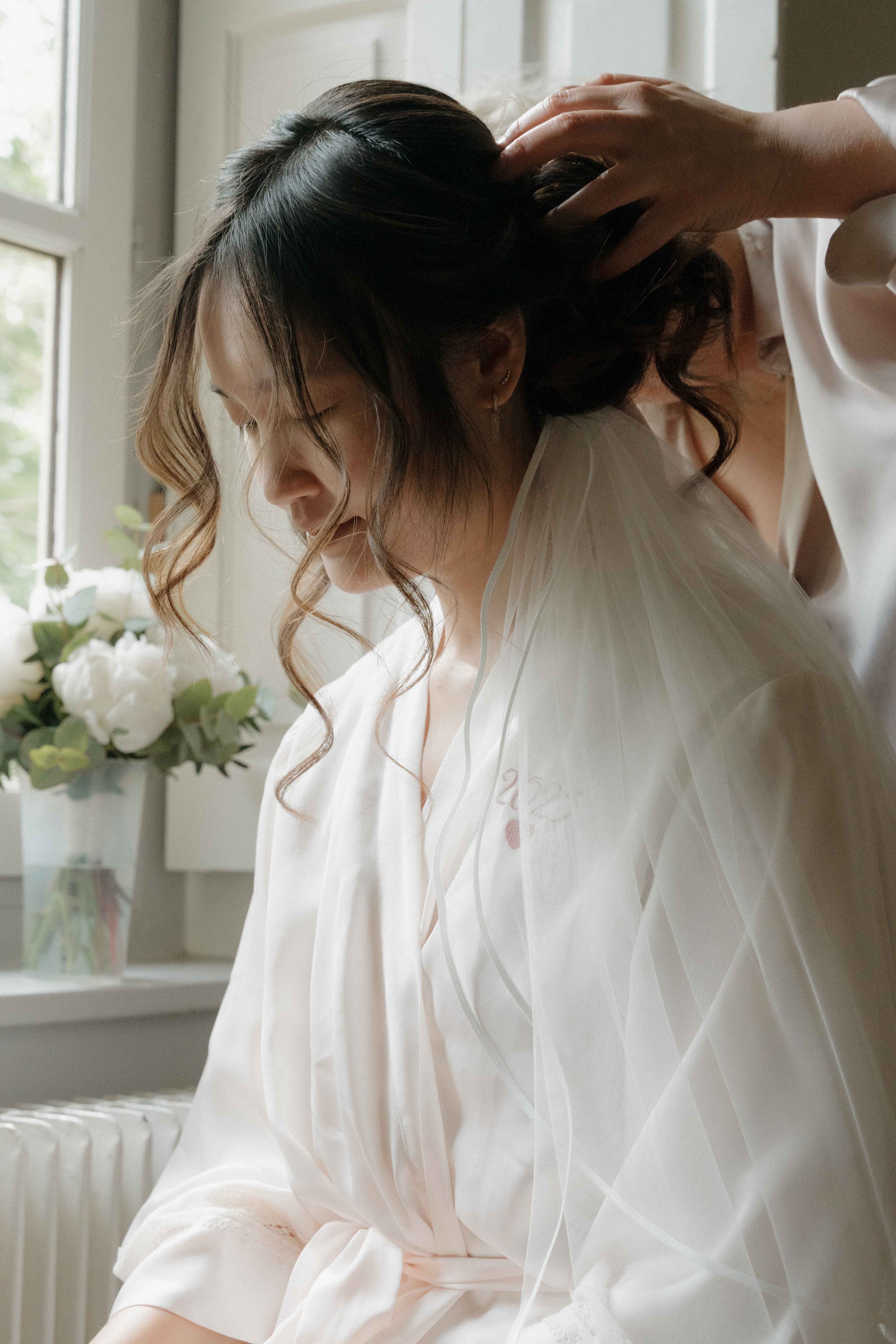 Une femme avec des cheveux bouclés subissant une coiffure ou maquillage, près d'une fenêtre avec une plante et des fleurs en arrière-plan. Domaine de tourieux à Savigny dans le beaujolais à Lyon. Mariage cérémonie laïque, photographe Lyon 