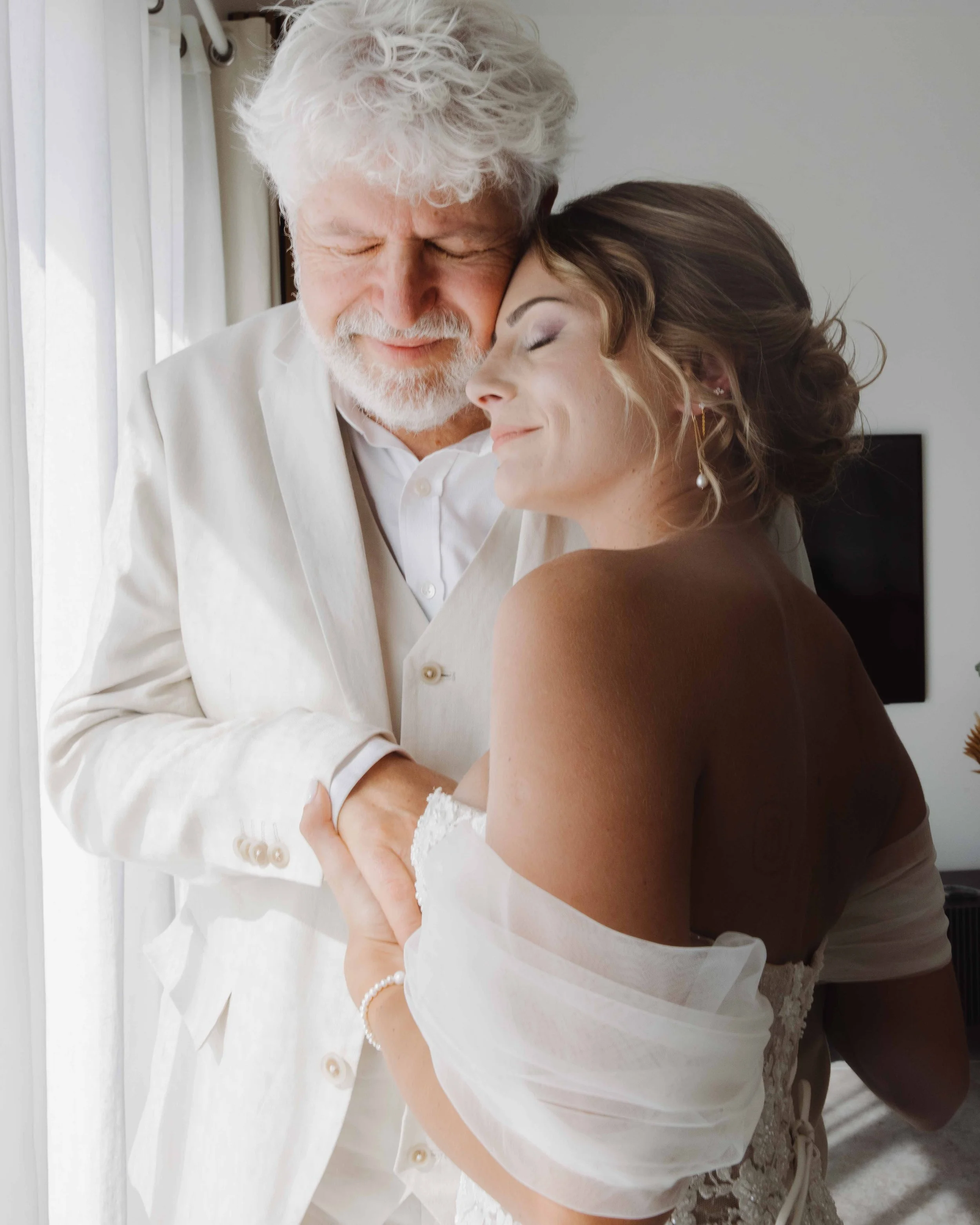 Un homme âgé et une jeune femme dans une étreinte chaleureuse, tous deux avec des expressions de bonheur et de tendresse, dans une pièce lumineuse. Photographe mariage Isère, ferme du rocher, cérémonie civil et laïque. Bordeaux et Lyon et destination