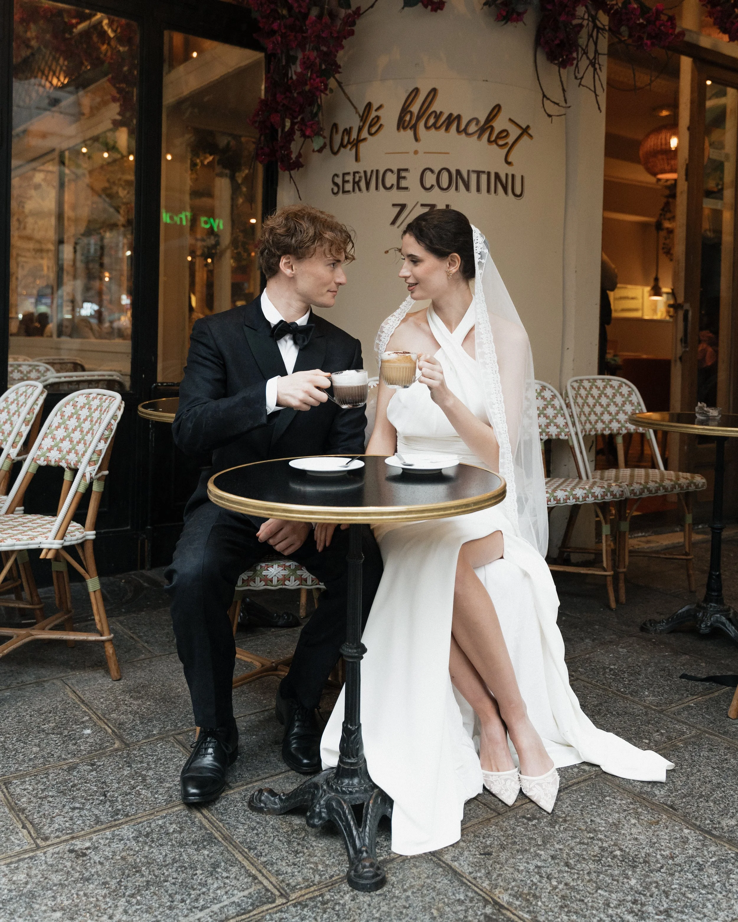 Un couple en mariage mangeant et buvant du café dans un café en plein air, avec une enseigne en arrière-plan qui indique 'Café Blanchet' et 'Service Continu'.