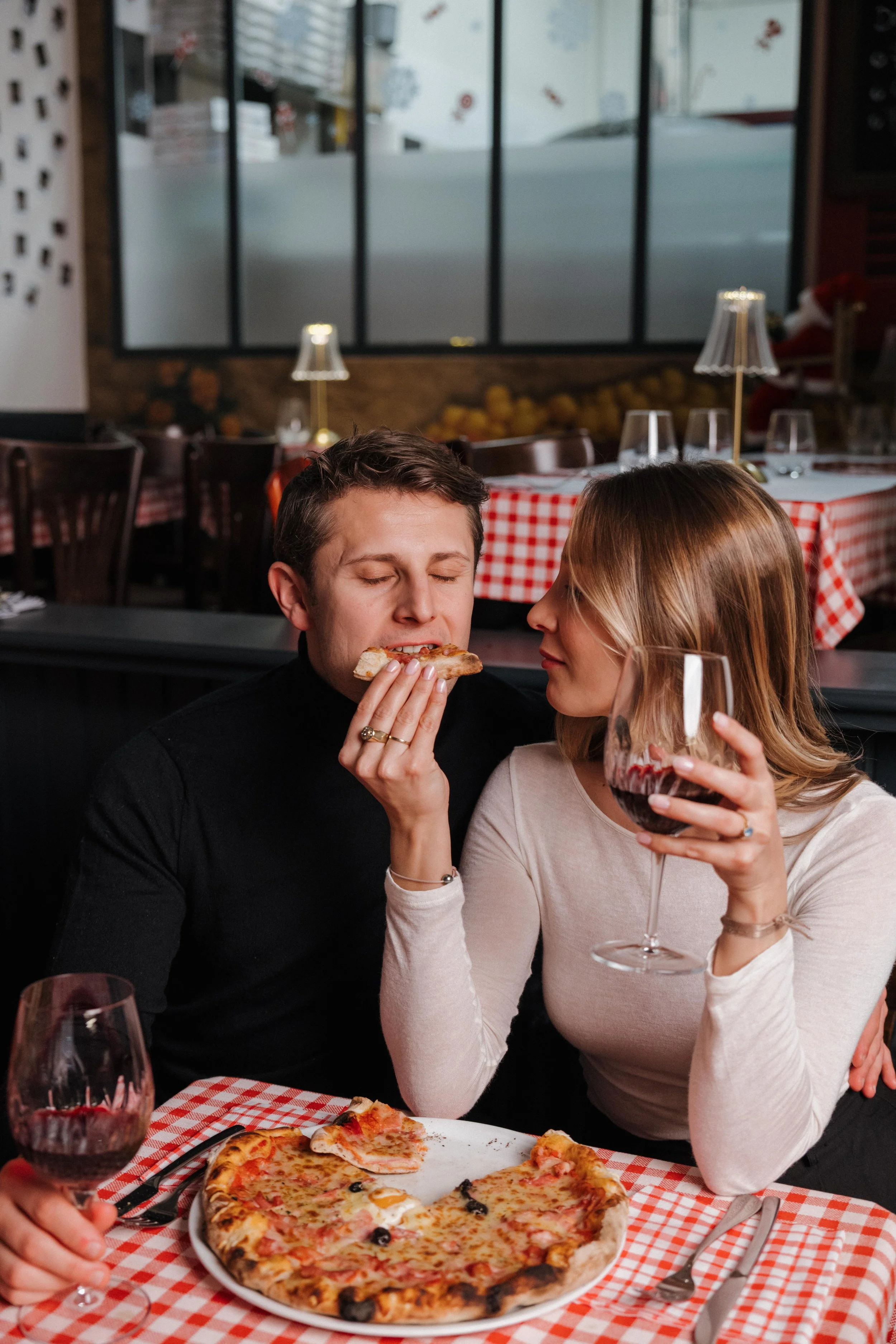 Un couple partageant une pizza au restaurant, la femme tient un verre de vin rouge, en arrière-plan décoration de Noël. session engagement couple Lyon 6eme, brotteaux, photographe mariage bordeaux et Lyon et destination wedding