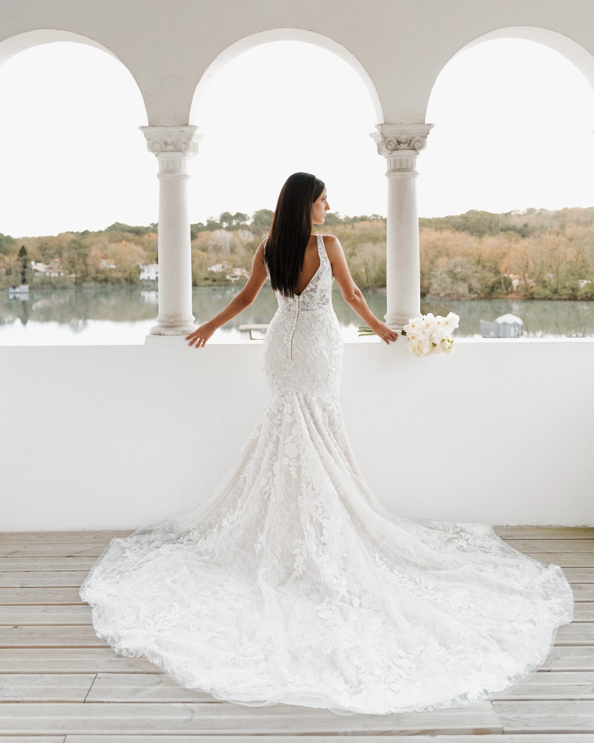 Une femme en robe de mariée blanche, vue de dos, tenant un bouquet blanc, devant une vue panoramique par une arche dans un environnement extérieur.  session femme mariées et robes haut de gamme, brindos, hôtel et lac,  photographe mariage bordeaux  