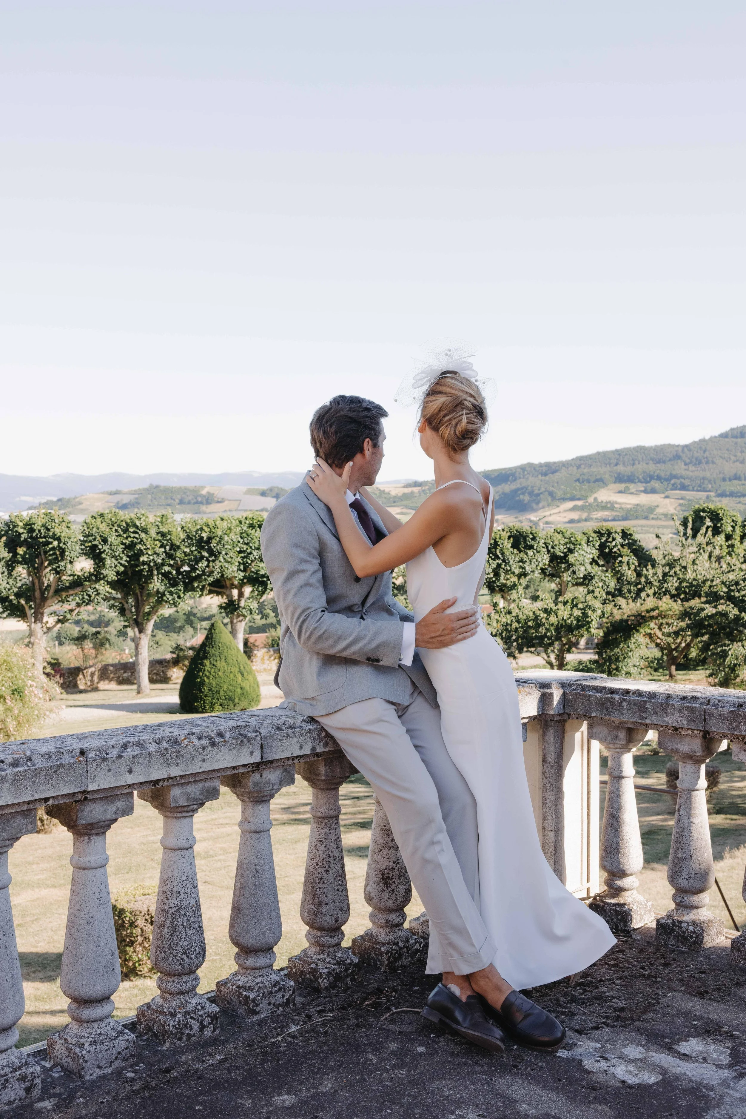 Un couple habillé élégamment, assis sur une balustrade en pierre, en pleine campagne avec des arbres et des collines en arrière-plan, lors d'une journée ensoleillée. Mariage au chateau de lachal dans le rhone alpes à côté de Lyon. Cérémonie civil 