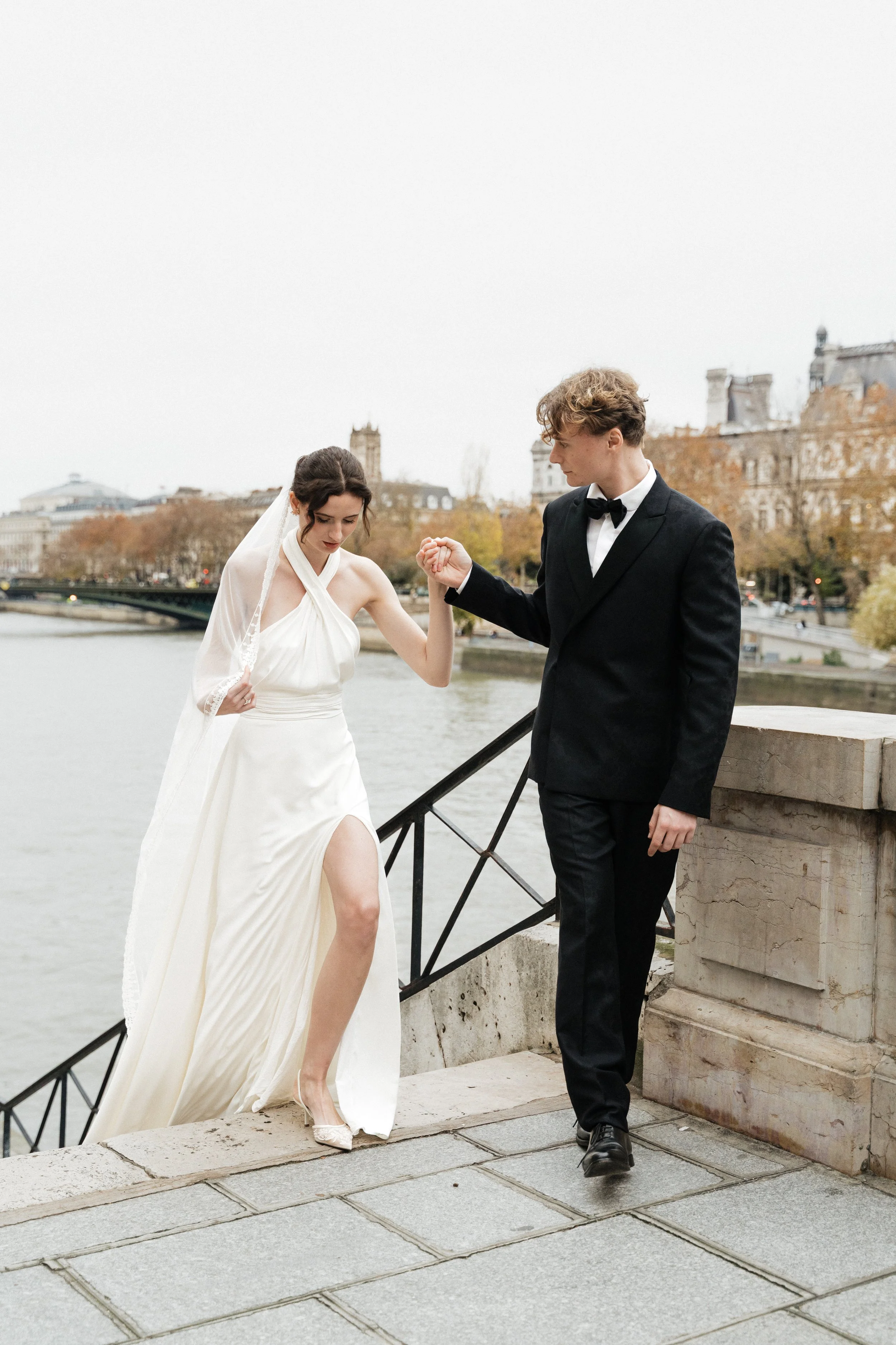 Un couple en mariage, la femme en robe blanche et le homme en costume noir, se tenant la main sur un pont au bord de la Seine à Paris. Session engagement paris, Louvre, mariage haut de gamme, destination wedding. Robe de mariées, madame a dit oui