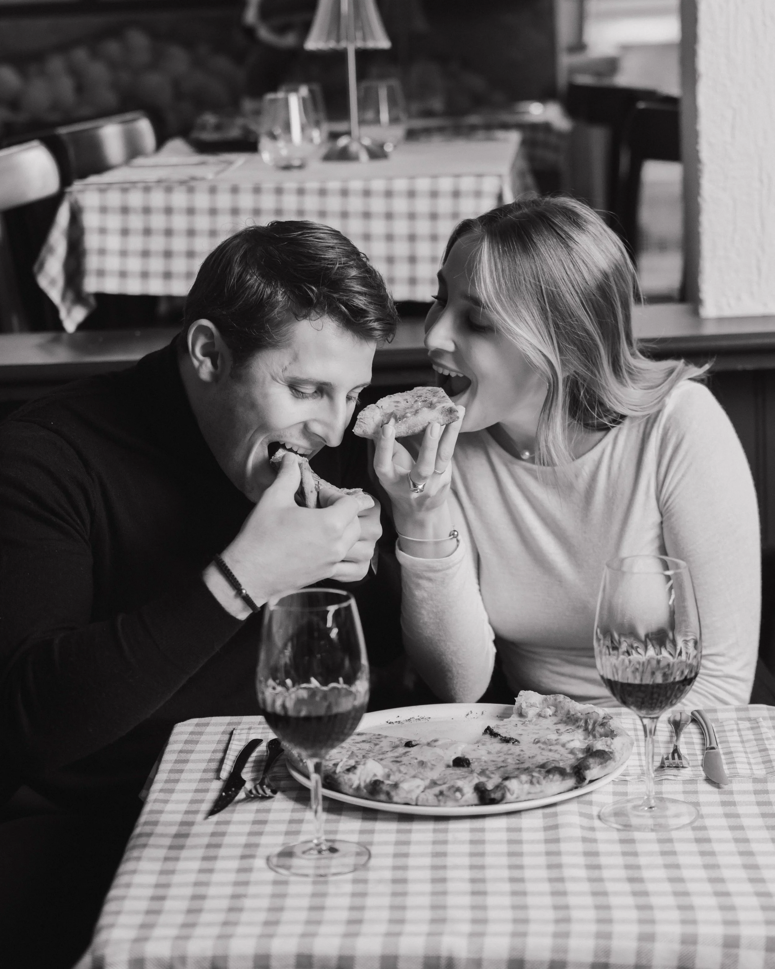 Un couple mangeant une pizza ensemble dans un restaurant, partageant un moment joyeux. La table a une nappe à carreaux et deux verres de vin. session engagement couple Lyon 6eme, brotteaux, photographe mariage bordeaux et Lyon et destination wedding