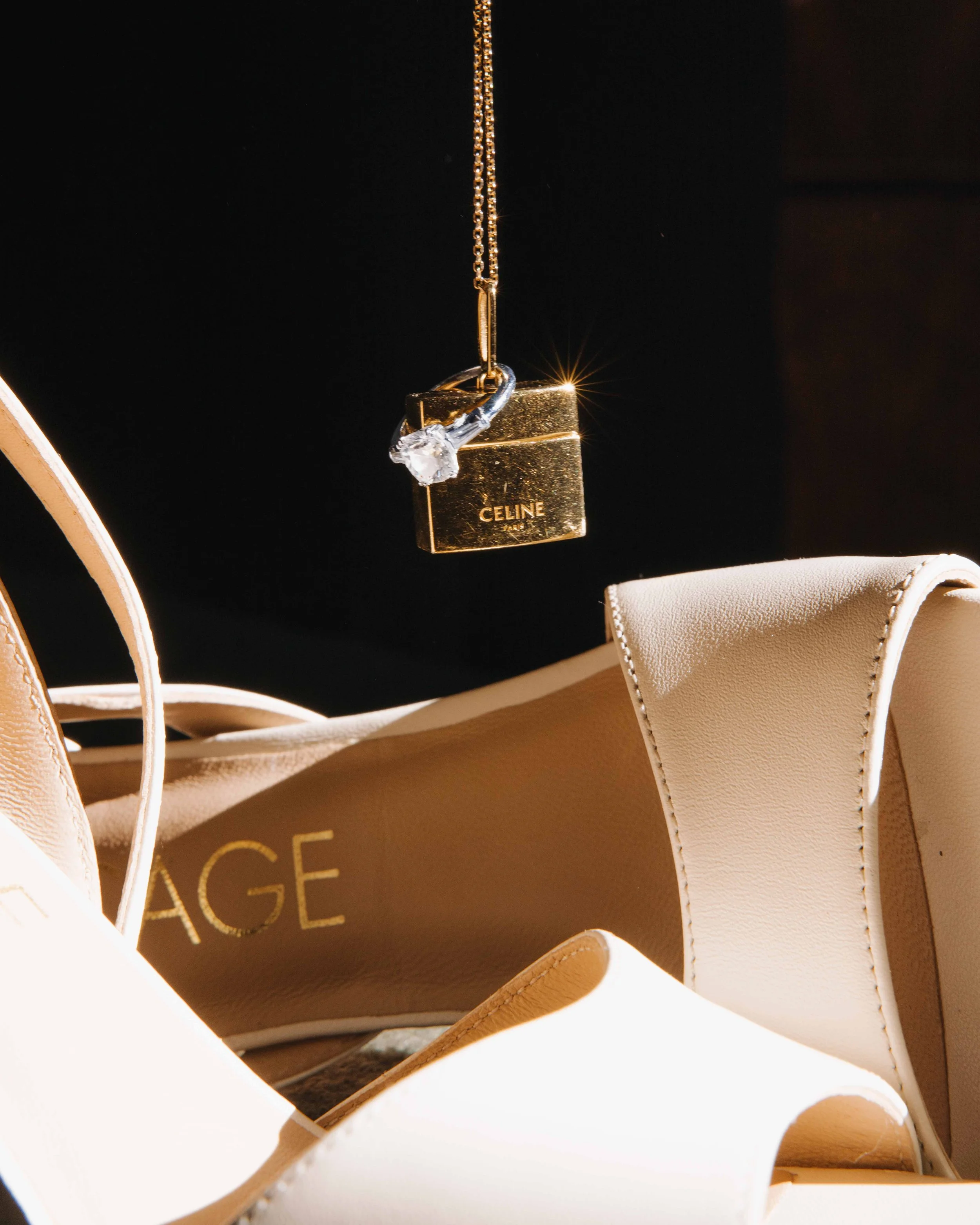 Une paire de chaussures à talons beiges, un sac de marque CELINE, et un collier avec un pendentif en forme de cube gold avec une pierre brillante, le tout contre un fond noir. Mariage au chateau de lachal dans le rhone alpes à côté de Lyon. 