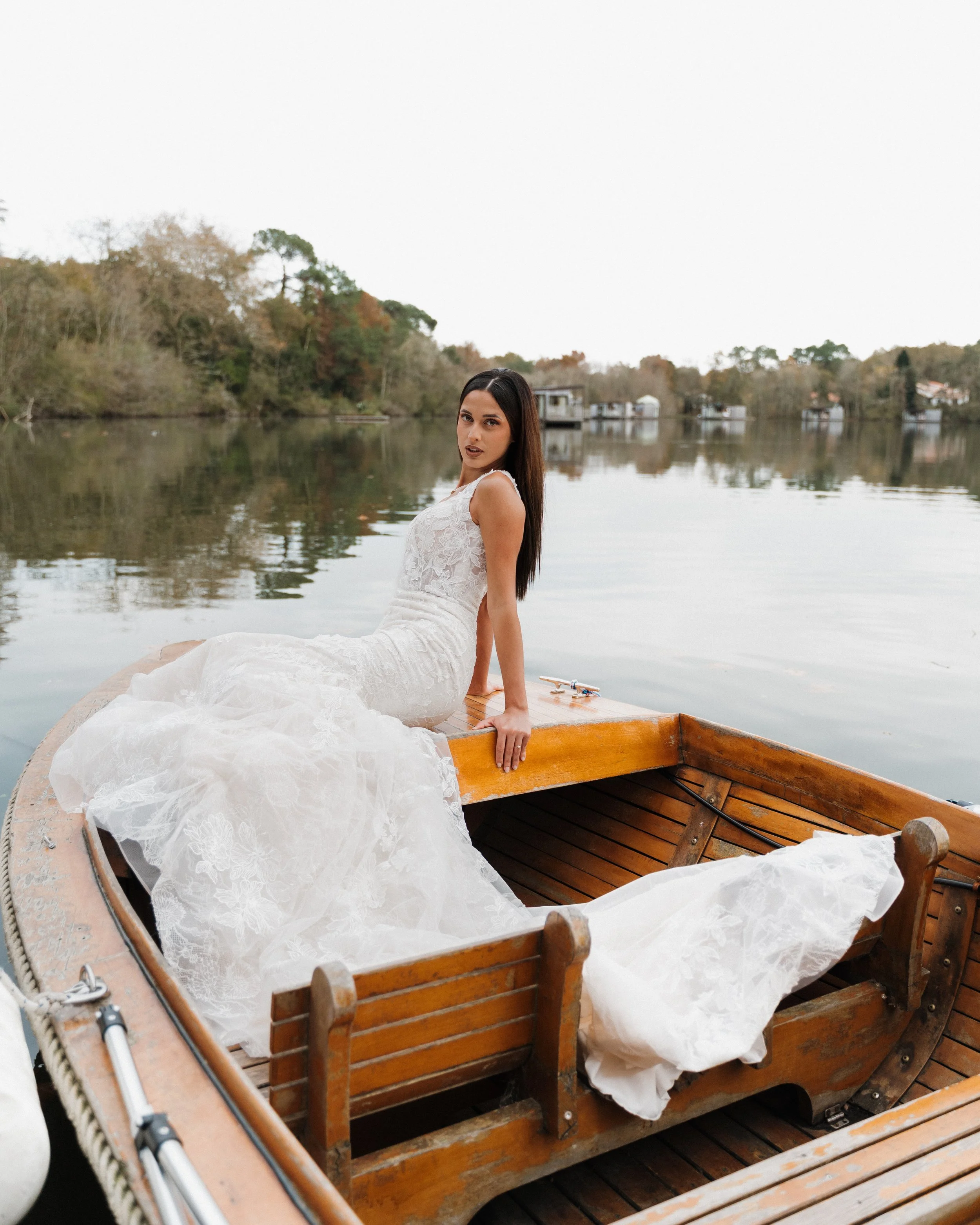 Une femme en robe de mariée blanche assise sur un bateau en bois au bord d'un lac, avec des arbres en arrière-plan.  session femme mariées et robes haut de gamme, brindos, hôtel et lac,  photographe mariage bordeaux et Lyon et destination wedding