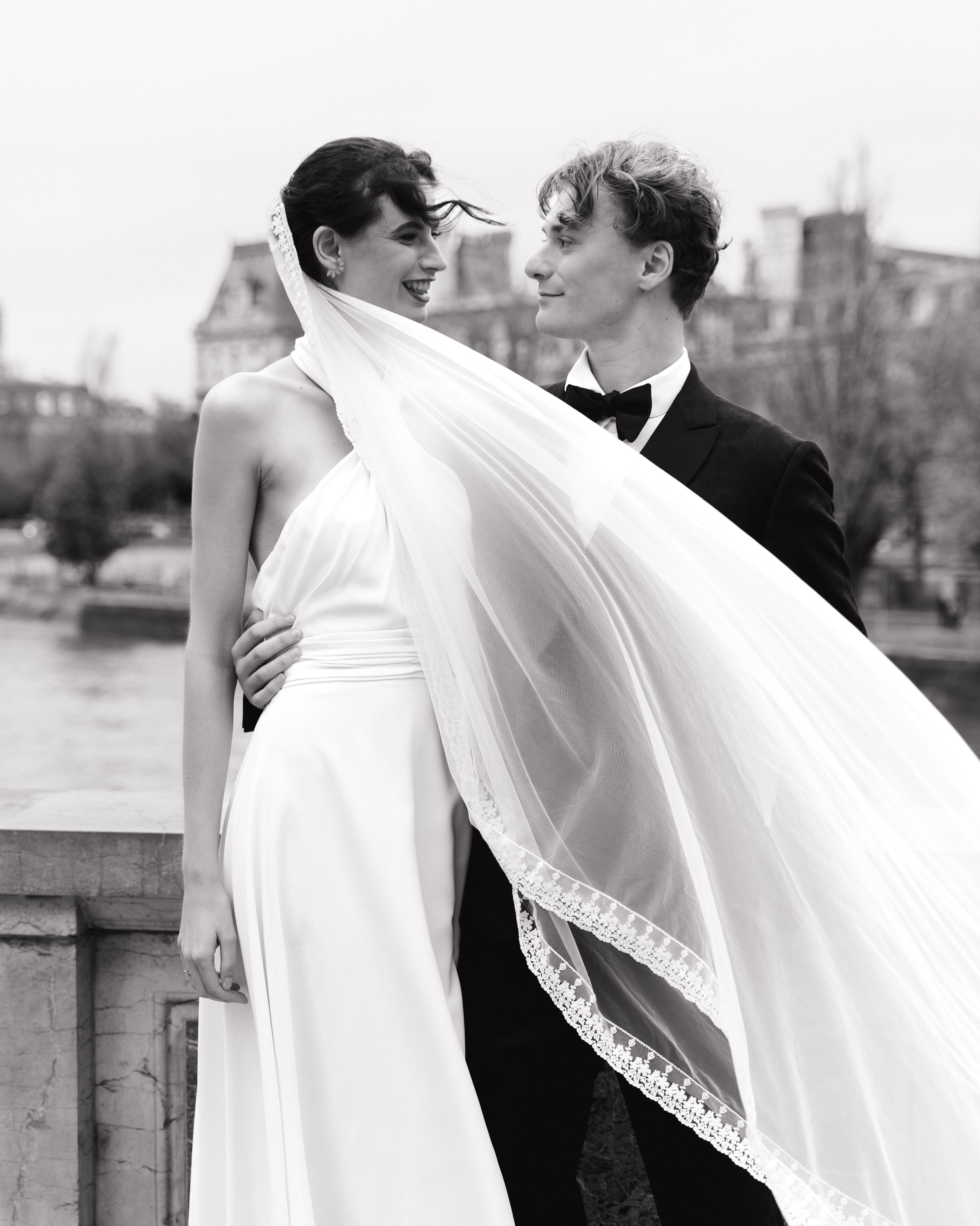 Un couple en robe de mariée et costume de marié lors d'une séance photo en extérieur, avec un bâtiment en arrière-plan. Session engagement paris, Louvre, mariage haut de gamme, destination wedding. Quai de seine, madame a dit oui